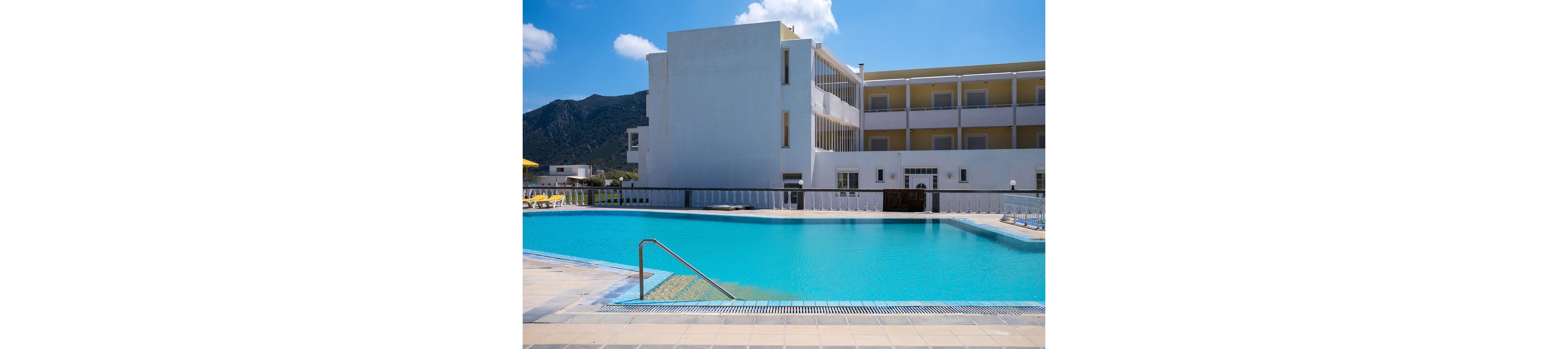 A large outdoor swimming pool sits in front of a modern white and yellow hotel building. Stainless steel handrails lead into the clear blue water, surrounded by tiled poolside decking. In the background, mountains rise under a bright blue sky with a few scattered clouds. The scene suggests a sunny, tranquil vacation setting at a resort property.