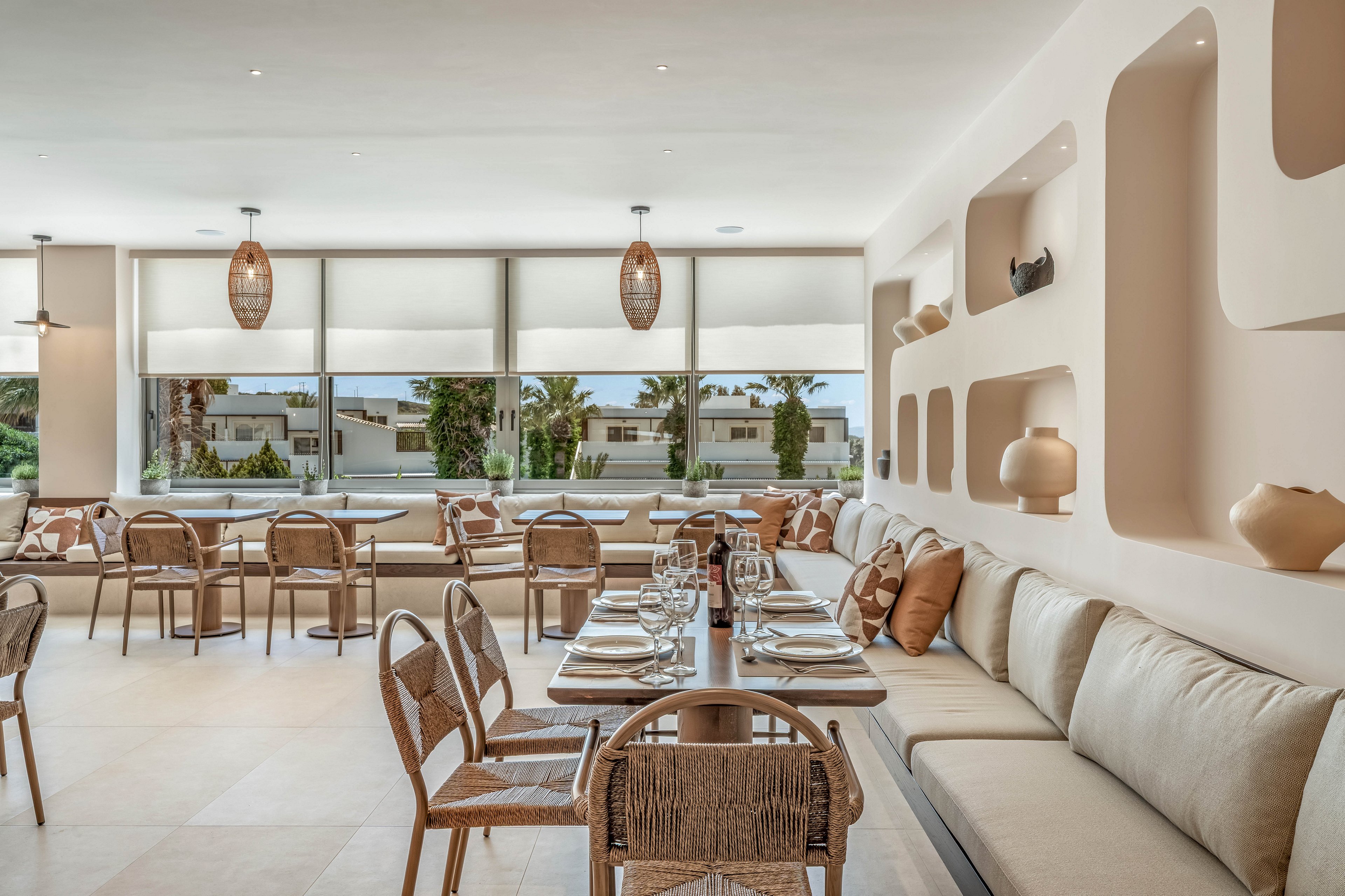 Contemporary restaurant dining area with neatly set tables and cushioned bench seating. Large windows with roller blinds let in natural light and reveal palm trees and low buildings outside. Built-in wall niches display decorative vases and lamps, complementing the beige and cream color palette. The atmosphere feels airy, relaxed, and inviting, ideal for casual or resort dining.