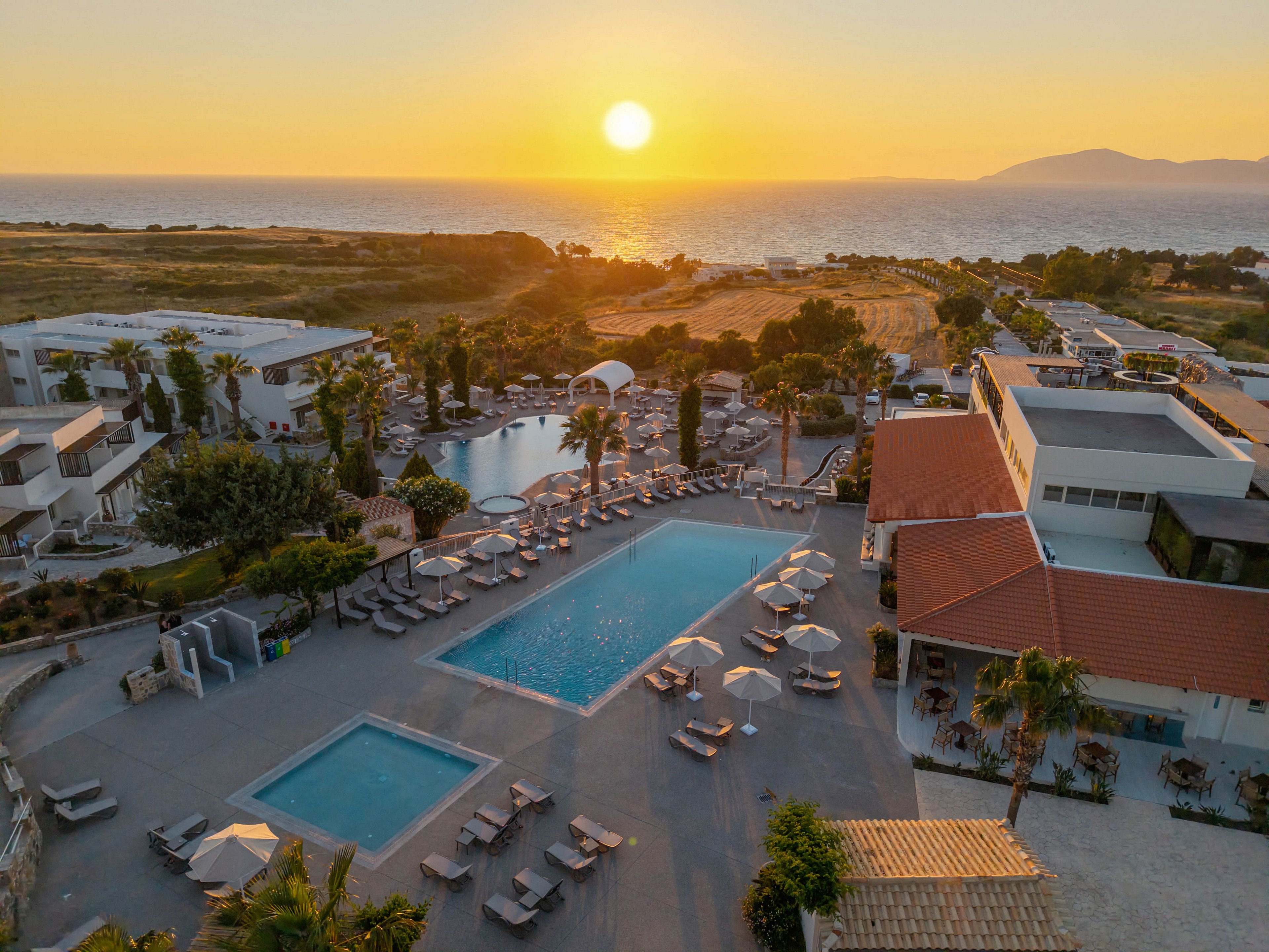 A large coastal resort is captured from above at sunset, featuring expansive swimming pools surrounded by sun loungers and umbrellas. The golden sun is low over the sea, casting warm light across the water and buildings. Palm trees and landscaped paths weave between the pools and accommodations, creating a relaxed vacation atmosphere. The distant coastline and hills frame the horizon, emphasizing the seaside location and tranquil mood.