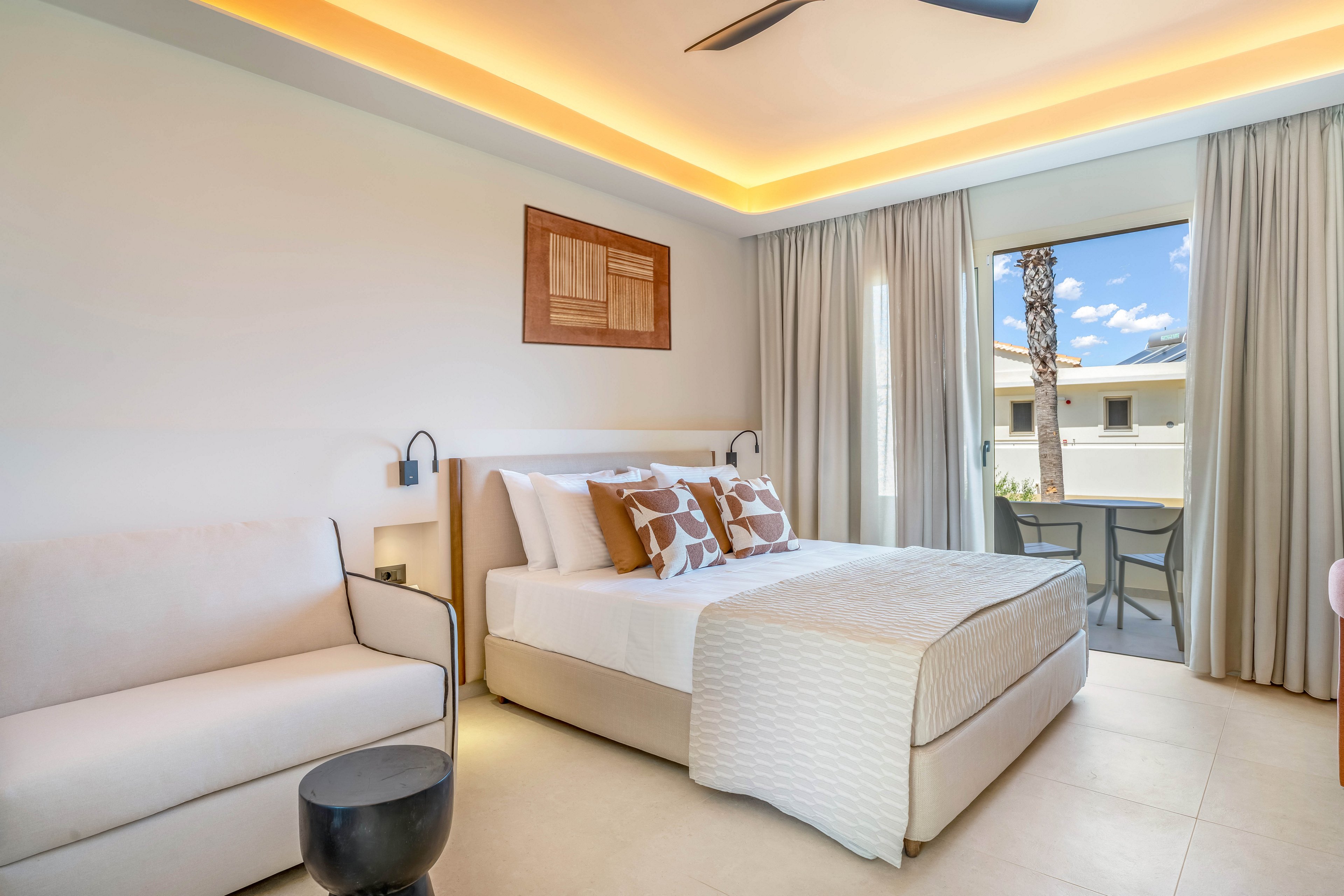 Contemporary hotel bedroom featuring a neatly made double bed with patterned pillows and a light quilt. The room includes a minimalist sofa, bedside tables, and wall-mounted reading lamps. Warm recessed ceiling lighting and neutral tones create a calm, inviting atmosphere. Large curtains frame a glass door that opens to an outdoor terrace with chairs and a view of palm trees and neighboring buildings.