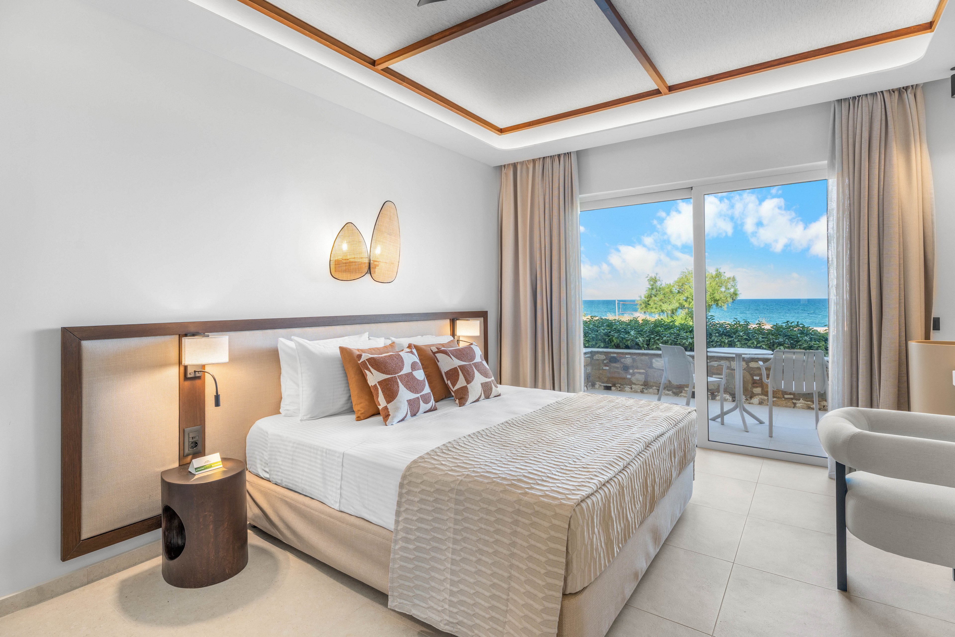 Modern hotel bedroom featuring a neatly made double bed with neutral bedding and patterned accent pillows. The room opens to a balcony through large sliding glass doors, revealing a clear view of the ocean and blue sky. Soft beige tones, wooden details, and contemporary lighting create a calm, upscale atmosphere. Outdoor chairs and greenery on the terrace emphasize the relaxing seaside setting.