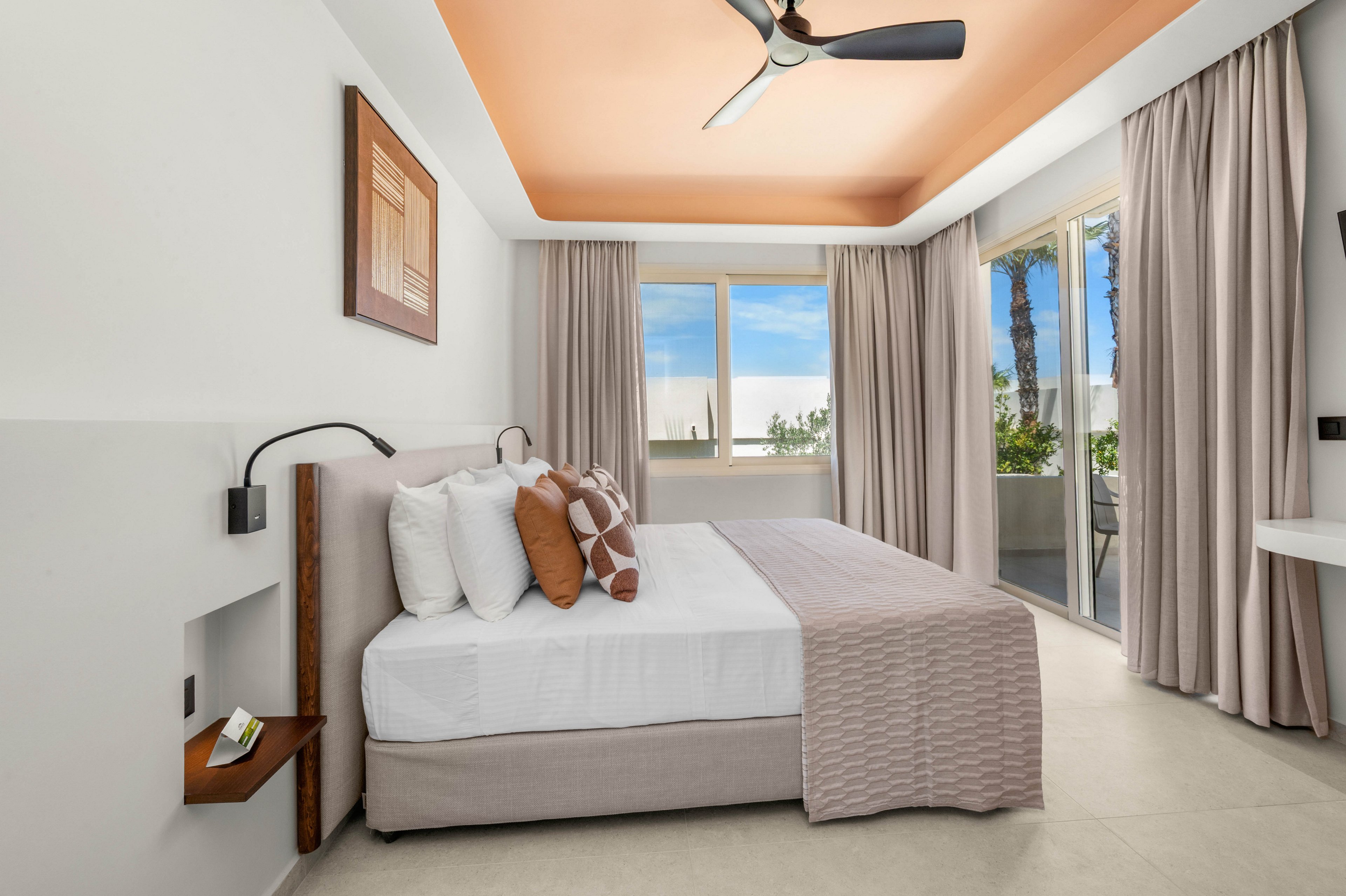 Contemporary hotel bedroom featuring a neatly made double bed with neutral bedding and decorative pillows. The room has minimalist furnishings, including wall-mounted bedside lights and a small built-in shelf. Large windows and a glass door open to a balcony with greenery and palm trees visible outside. Soft beige and white tones with a warm peach ceiling create a calm, inviting atmosphere.