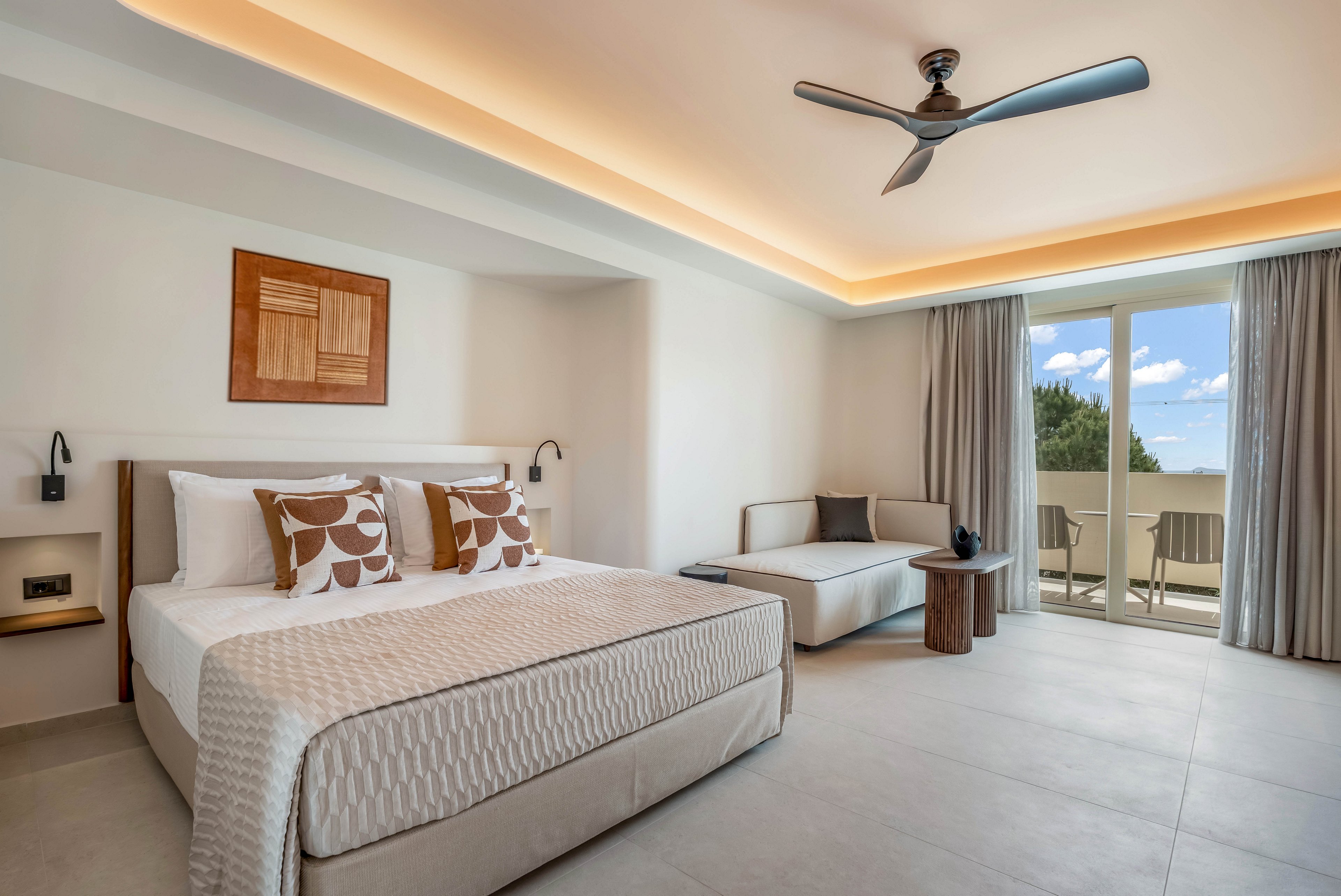 Spacious contemporary hotel room featuring a large bed with patterned cushions and neutral bedding. The space includes a sleek sofa, small wooden side table, and ceiling fan. Warm recessed lighting highlights the minimalist decor and clean lines. Floor-to-ceiling glass doors open to a balcony with outdoor seating and a view of trees and blue sky.