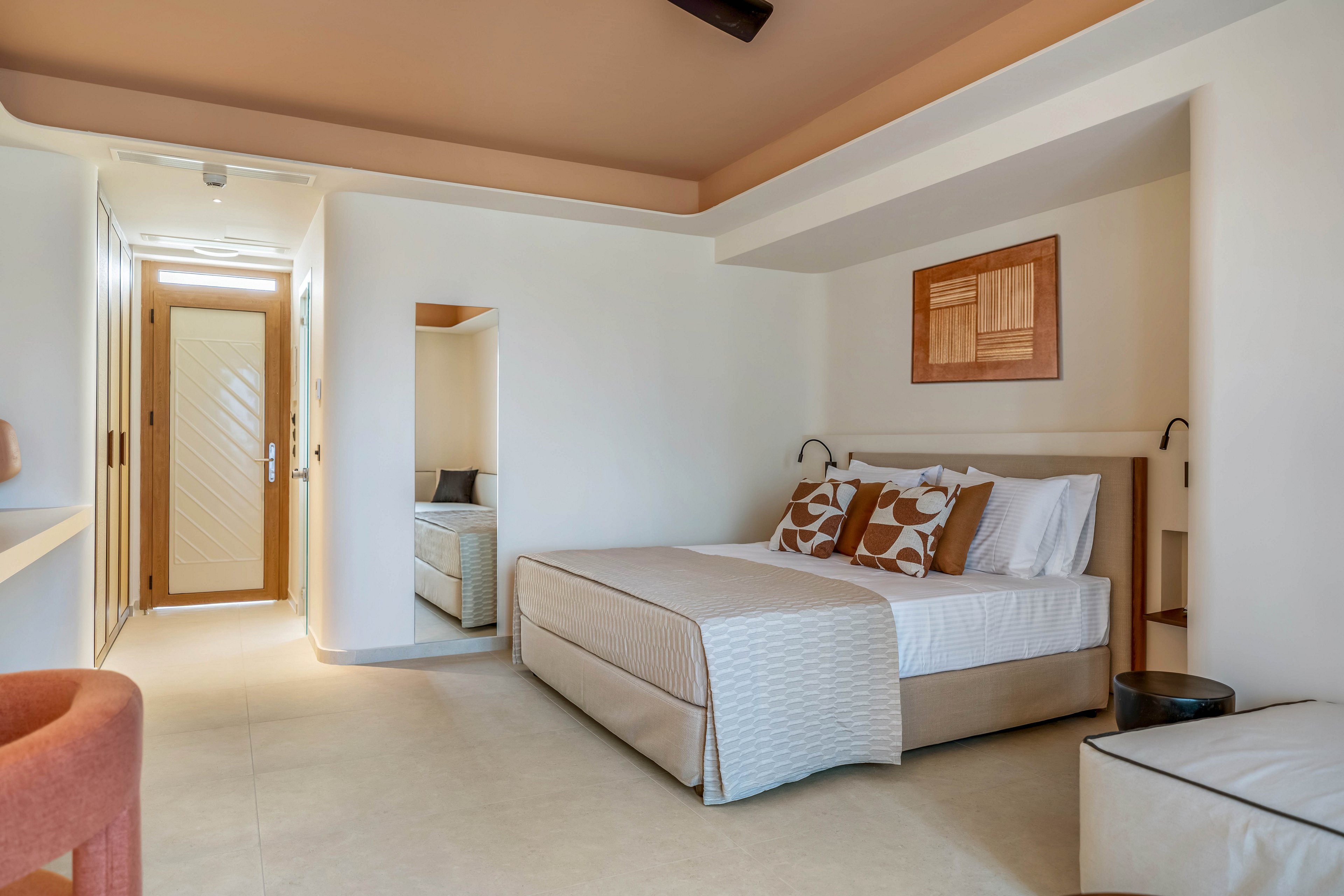 A contemporary hotel bedroom features a neatly made double bed with patterned cushions and a light quilt. The room is decorated in warm beige and terracotta tones, with a matching ceiling and upholstered chair. A wooden artwork hangs above the bed, and built-in reading lamps flank the headboard. In the background, a hallway leads to a door and a small adjoining seating or sleeping nook, enhancing the spacious, airy feel.