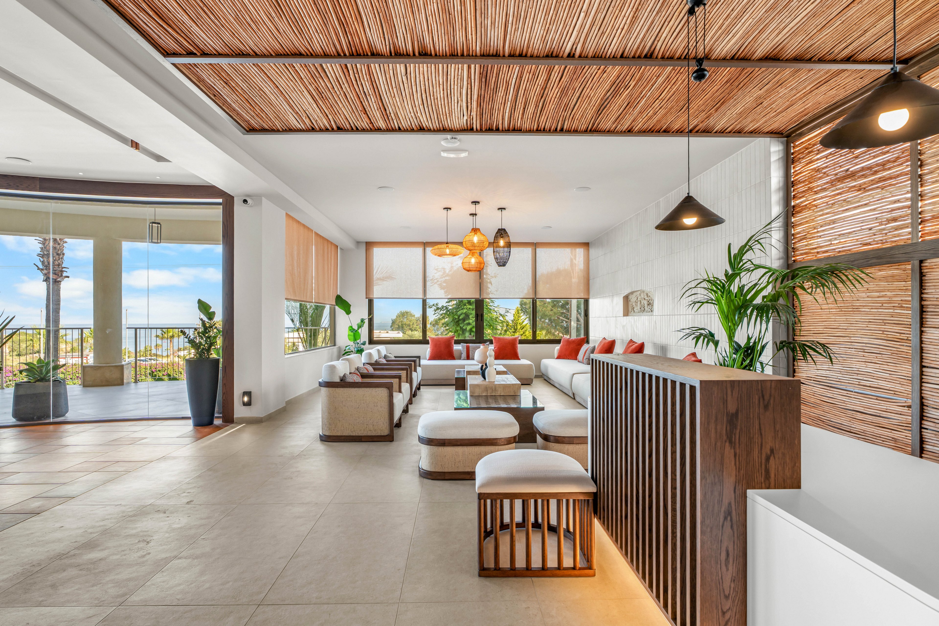 Spacious contemporary lounge area features low white sofas and cushioned stools arranged around minimalist tables. Large windows and sliding glass doors open to a balcony with ocean or coastal views. Warm wooden ceiling slats, woven shades, and pendant lights create a relaxed, resort-like atmosphere. Green potted plants and neutral-toned decor enhance the airy, inviting interior space.