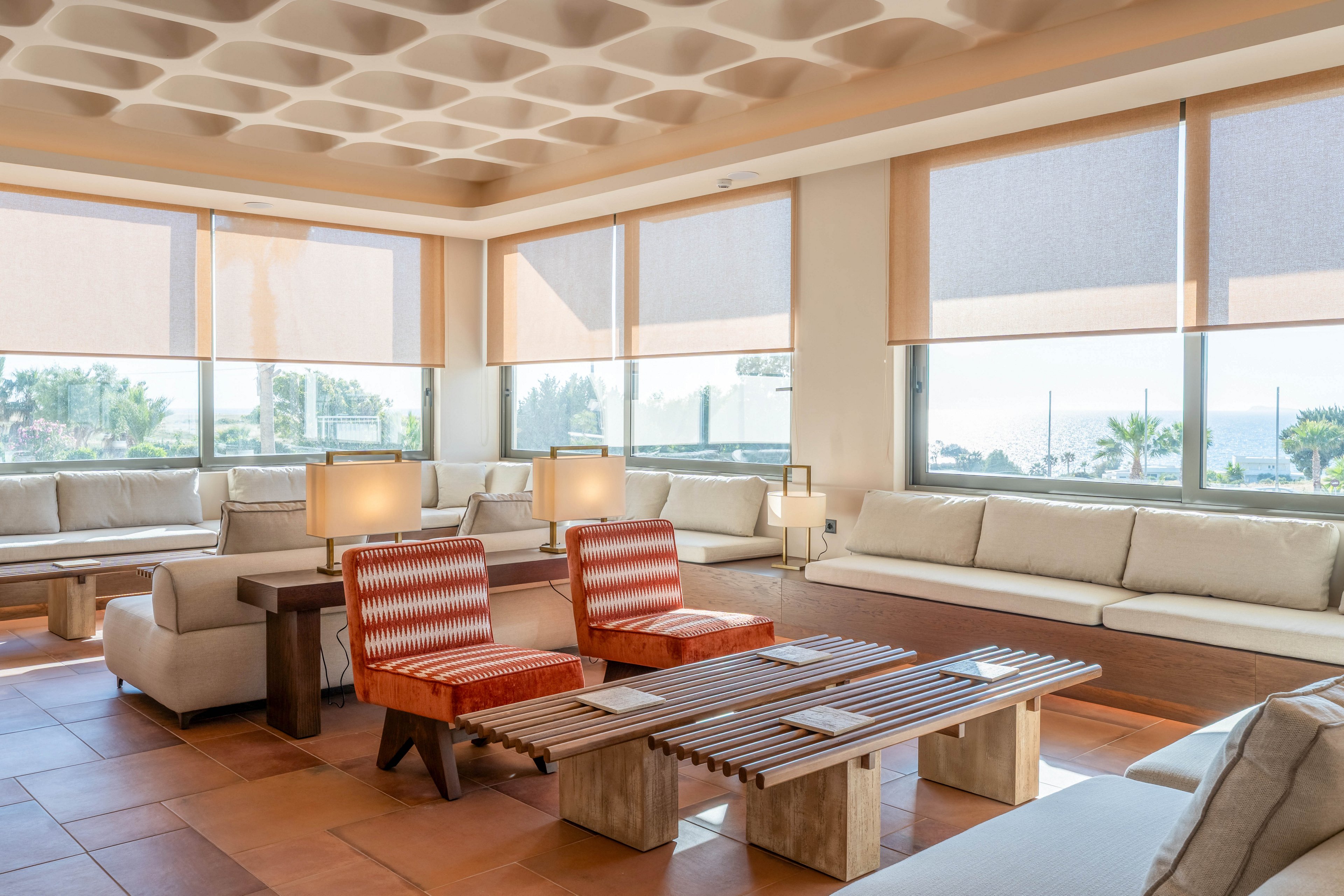 A spacious modern lounge features long white sofas and two patterned armchairs around a slatted wooden coffee table. Large windows with roller shades let in abundant natural light and reveal a glimpse of palm trees and the sea outside. Warm neutral tones dominate the tiled floor, furniture, and ceiling design, creating a relaxed yet upscale atmosphere. Table lamps and clean lines emphasize a contemporary, comfortable interior setting.