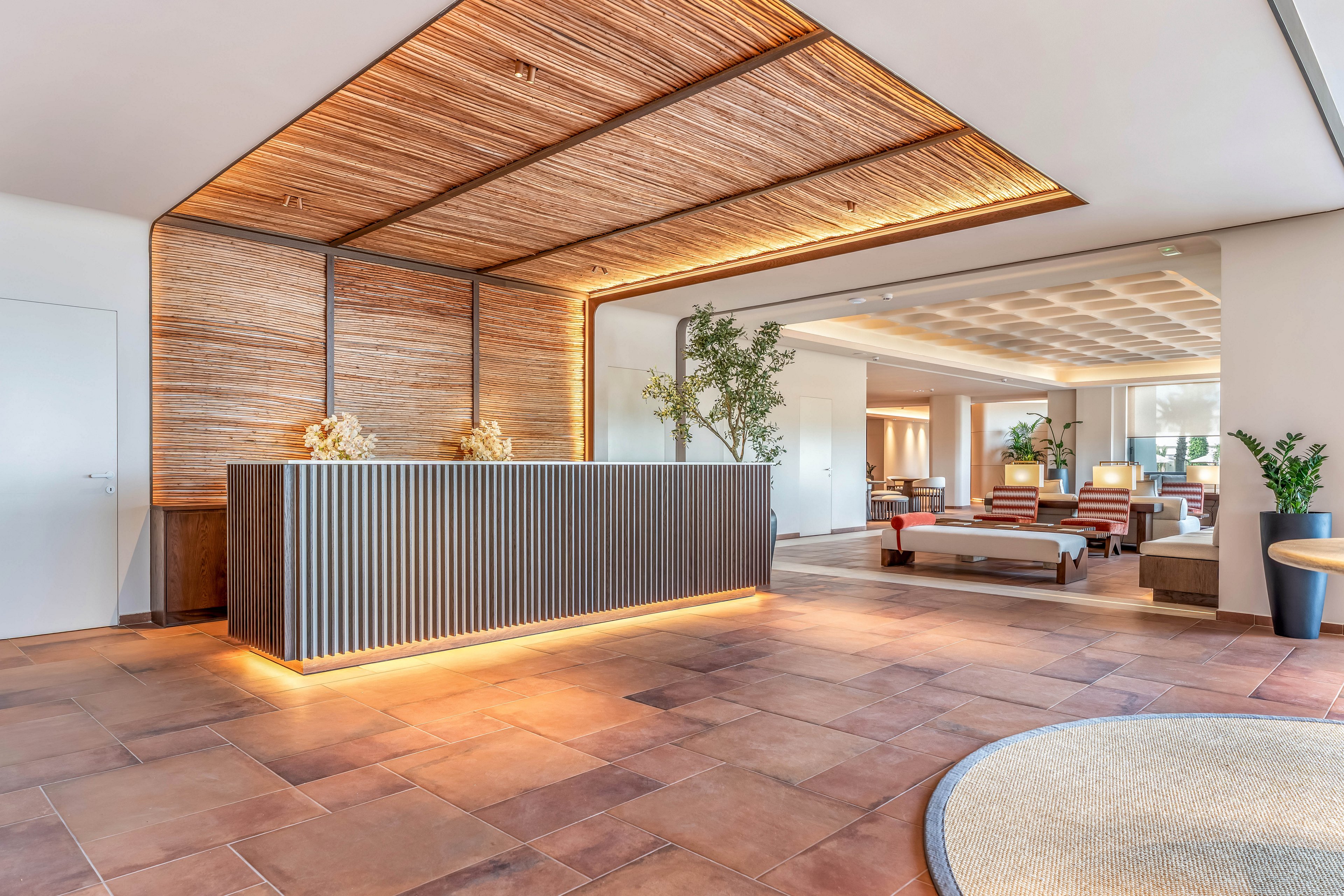 A contemporary hotel lobby features a sleek reception desk with vertical slats and warm underlighting. The space is designed with natural materials, including a wood-paneled ceiling and terracotta-style floor tiles. Comfortable seating areas with sofas and armchairs extend into the background, creating an inviting lounge atmosphere. Potted plants and soft, indirect lighting contribute to a calm, welcoming environment.
