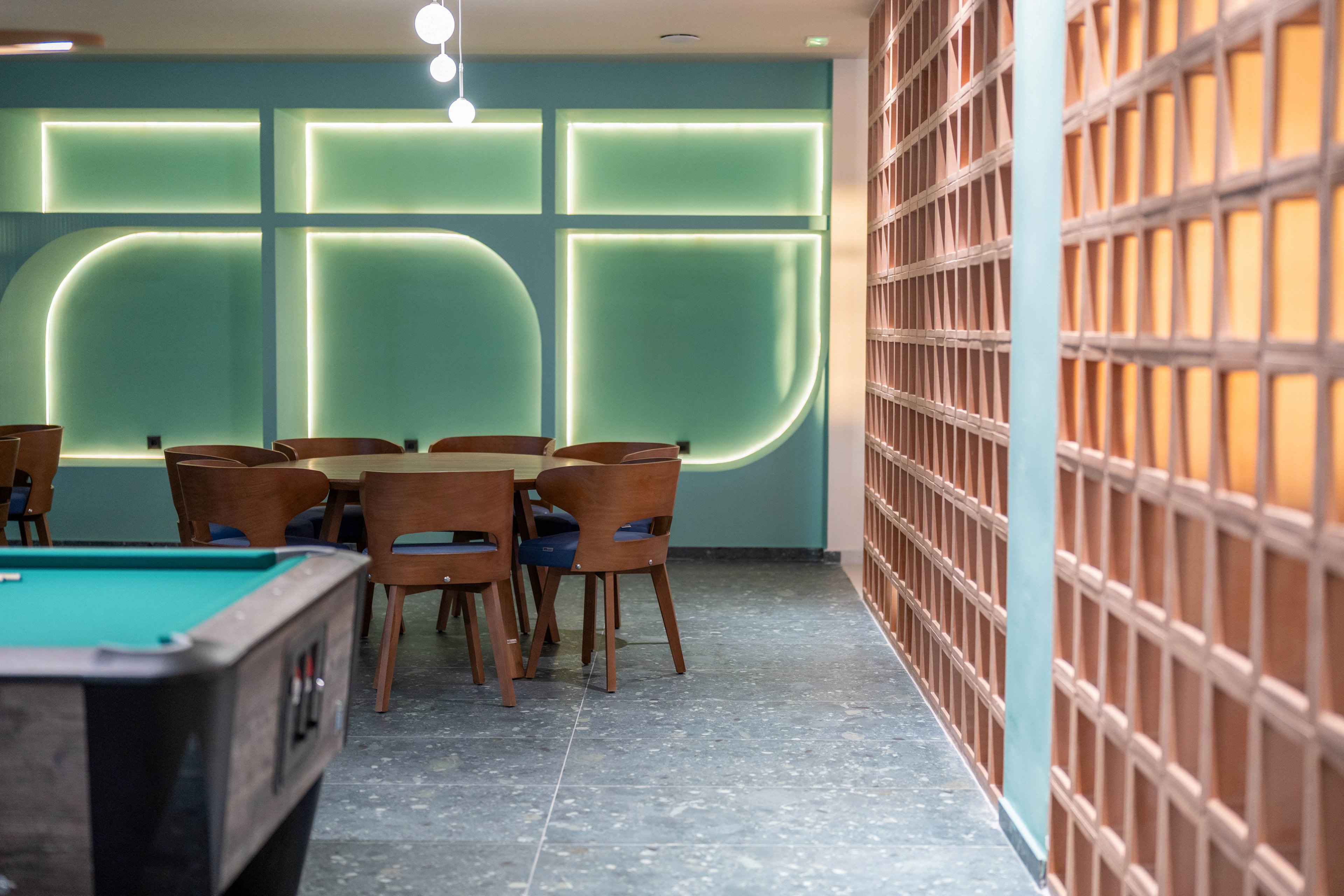Contemporary lounge interior featuring a pool table in the foreground and a wooden dining table with chairs in the background. The setting includes a decorative wall with geometric neon-style lighting that creates a stylish focal point. Warm wood tones contrast with cool teal walls and flooring, giving the space a cozy yet modern feel. The room appears quiet and empty, suggesting a relaxed social or recreational area.