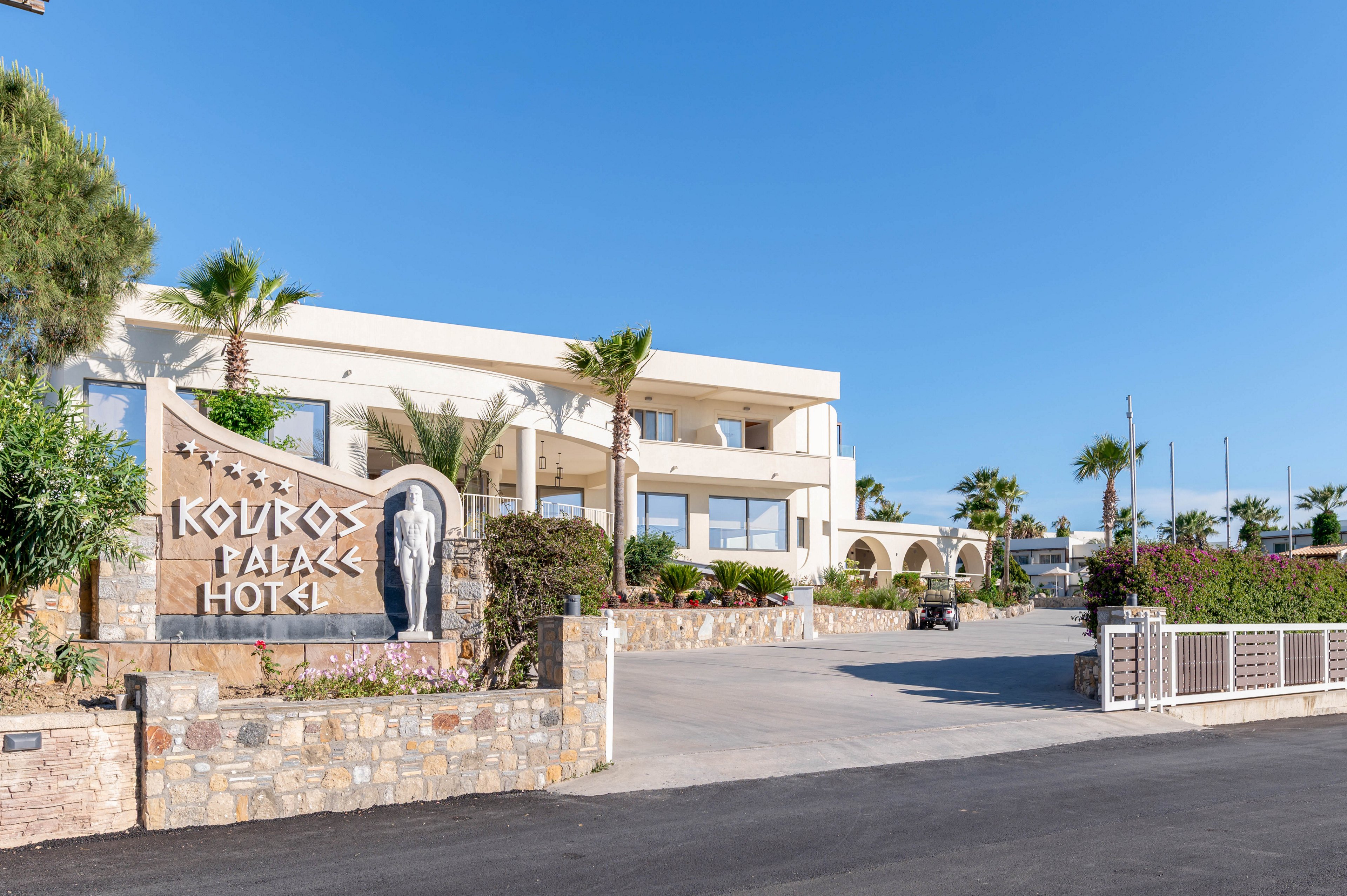 A bright exterior view of the Kouros Palace Hotel shows a modern, cream-colored building under a clear blue sky. The main entrance is framed by palm trees, stone walls, and a wide driveway leading up to the hotel. A large decorative sign with the hotel name and a sculpted figure stands prominently in the foreground. The scene conveys a sunny, welcoming atmosphere typical of a Mediterranean resort destination.