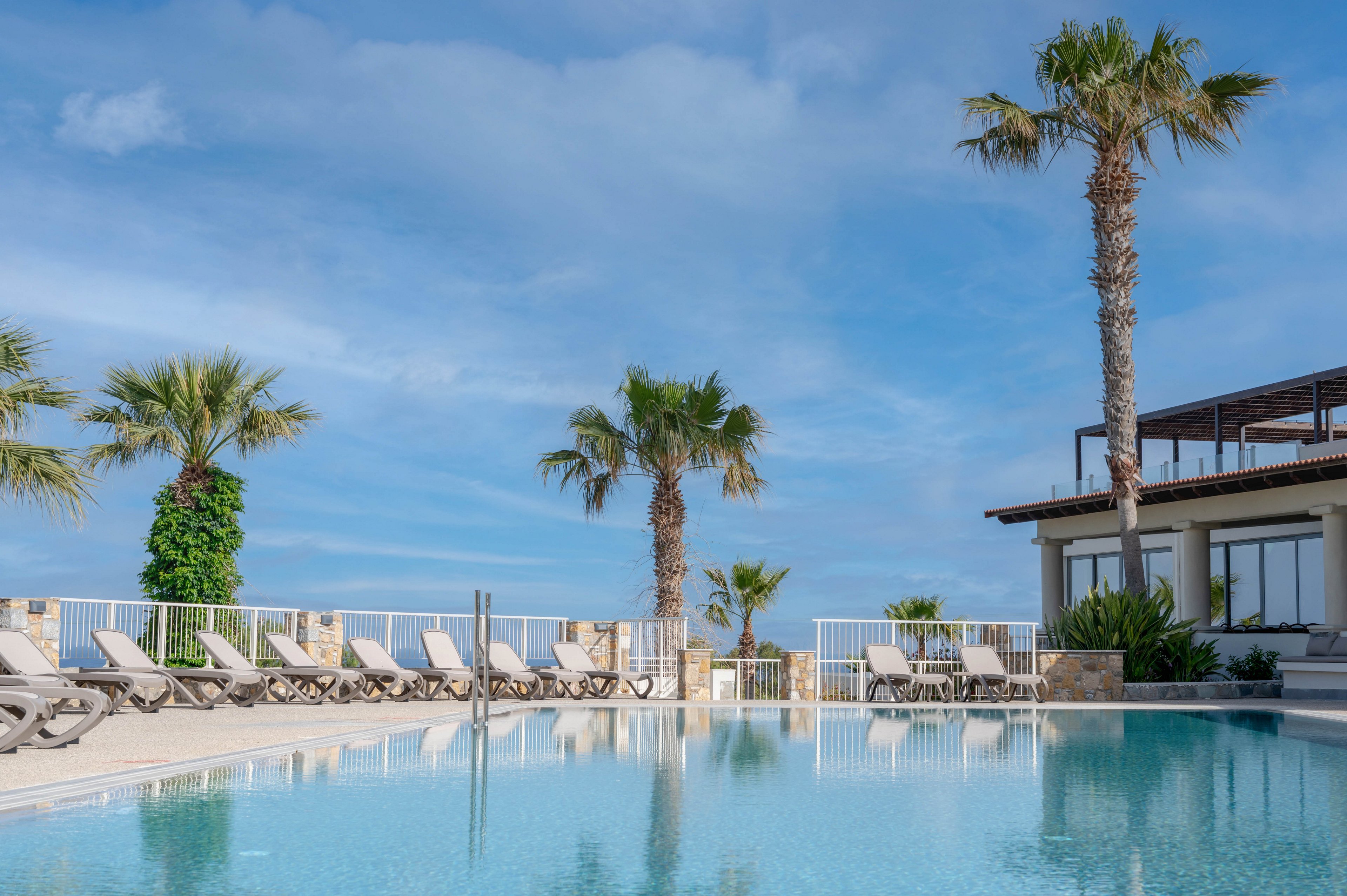 An inviting resort swimming pool reflects the clear blue sky and surrounding palm trees. Numerous empty sun loungers line the poolside terrace, suggesting a tranquil and uncrowded atmosphere. A modern building with large windows and balconies stands to the right, overlooking the water. The overall scene conveys a sunny, relaxing vacation setting in a warm coastal climate.