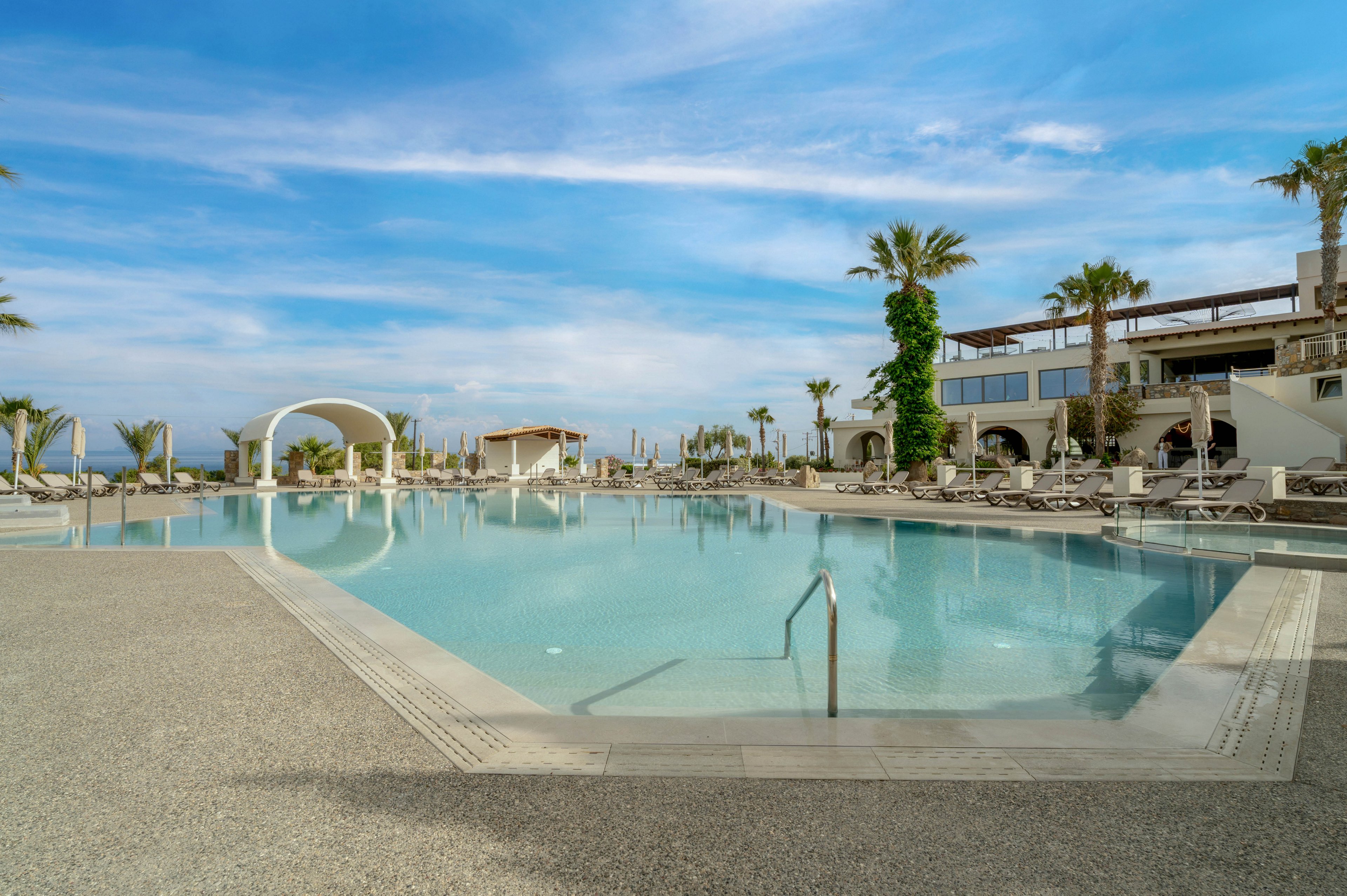 A large, tranquil infinity pool stretches across the foreground of a modern seaside resort. Sun loungers and umbrellas line the poolside, suggesting a relaxing vacation setting. Palm trees and contemporary white arches frame the scene under a bright blue sky with scattered clouds. The calm water and spacious deck create an inviting, serene atmosphere for guests.