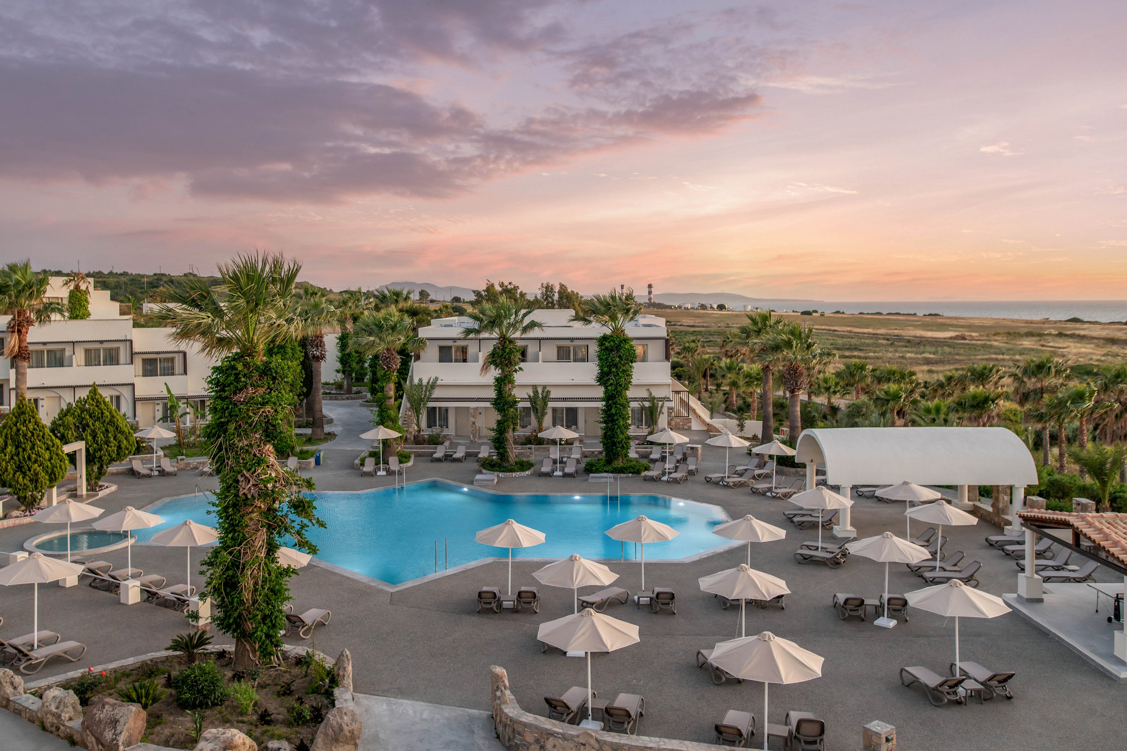 A large resort swimming pool is surrounded by white umbrellas and lounge chairs in a spacious courtyard. Modern low-rise buildings with balconies and tall palm trees frame the pool area. The sky is filled with soft pastel colors as the sun sets over distant fields and coastline. The scene feels calm and inviting, suggesting a relaxing vacation destination.