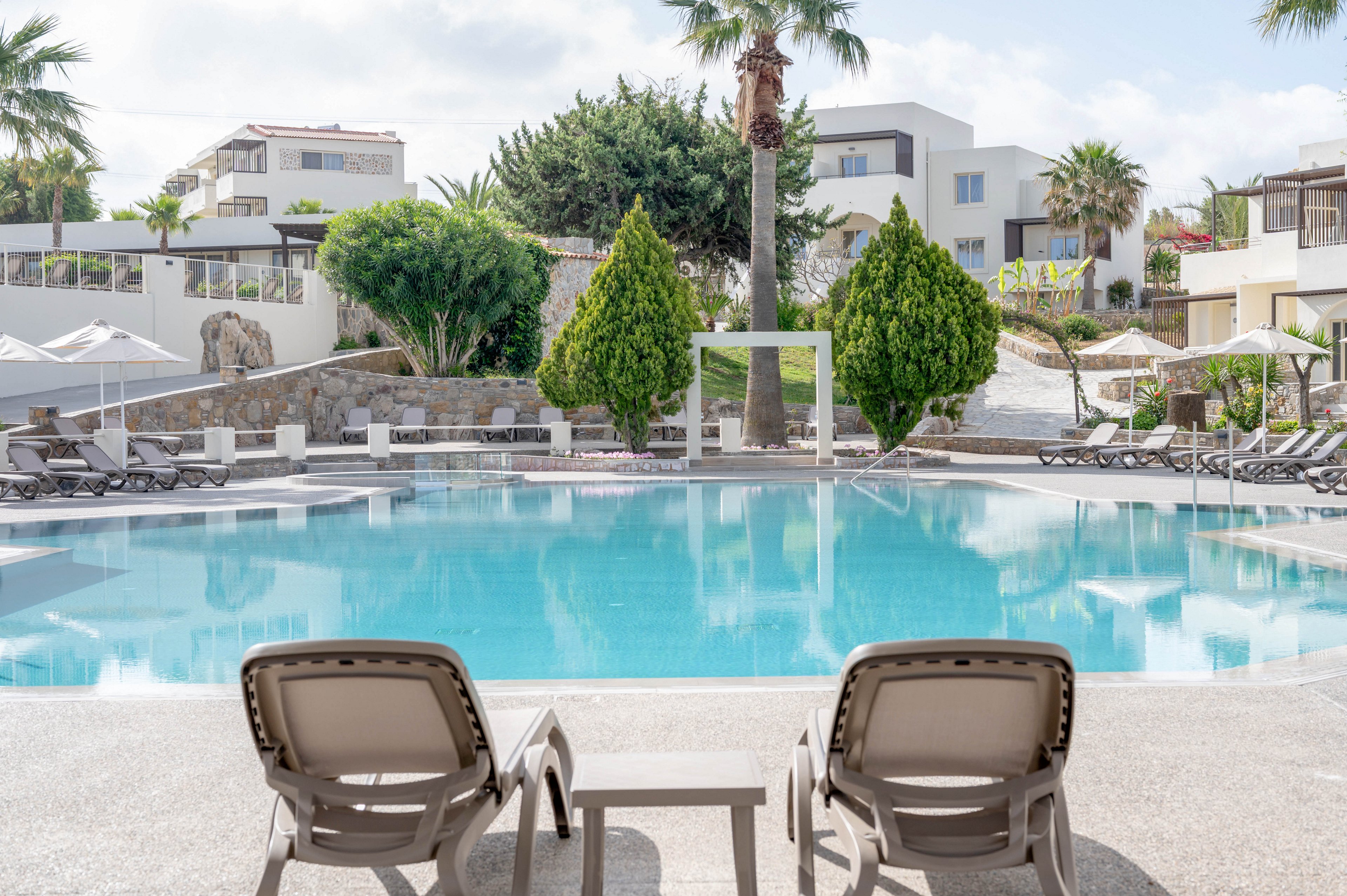 A tranquil resort swimming pool is framed by two empty sun loungers and a small table in the foreground. The pool is surrounded by neatly arranged deck chairs and umbrellas, suggesting a relaxing vacation setting. Modern white buildings and lush green trees rise behind the pool, creating a clean and inviting atmosphere. Soft daylight and calm water reflections contribute to a peaceful, leisurely mood.