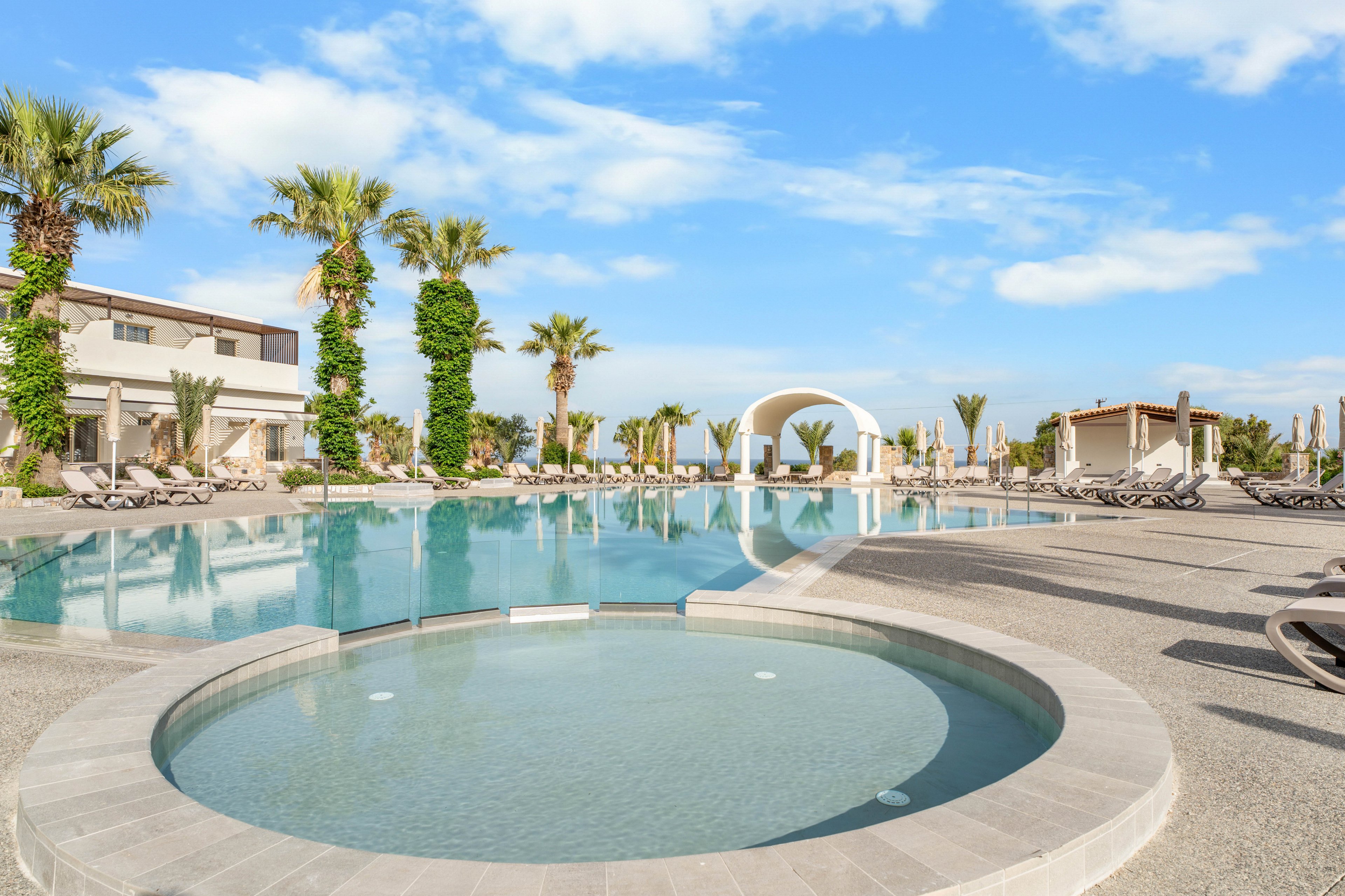 A luxurious resort features a large outdoor swimming pool with a circular spa pool in the foreground. Palm trees and modern buildings line the poolside, creating a tropical vacation atmosphere. Sun loungers and umbrellas are neatly arranged along the water’s edge, suggesting a relaxing holiday setting. The sky is bright with scattered clouds, reflecting softly on the calm pool surface.