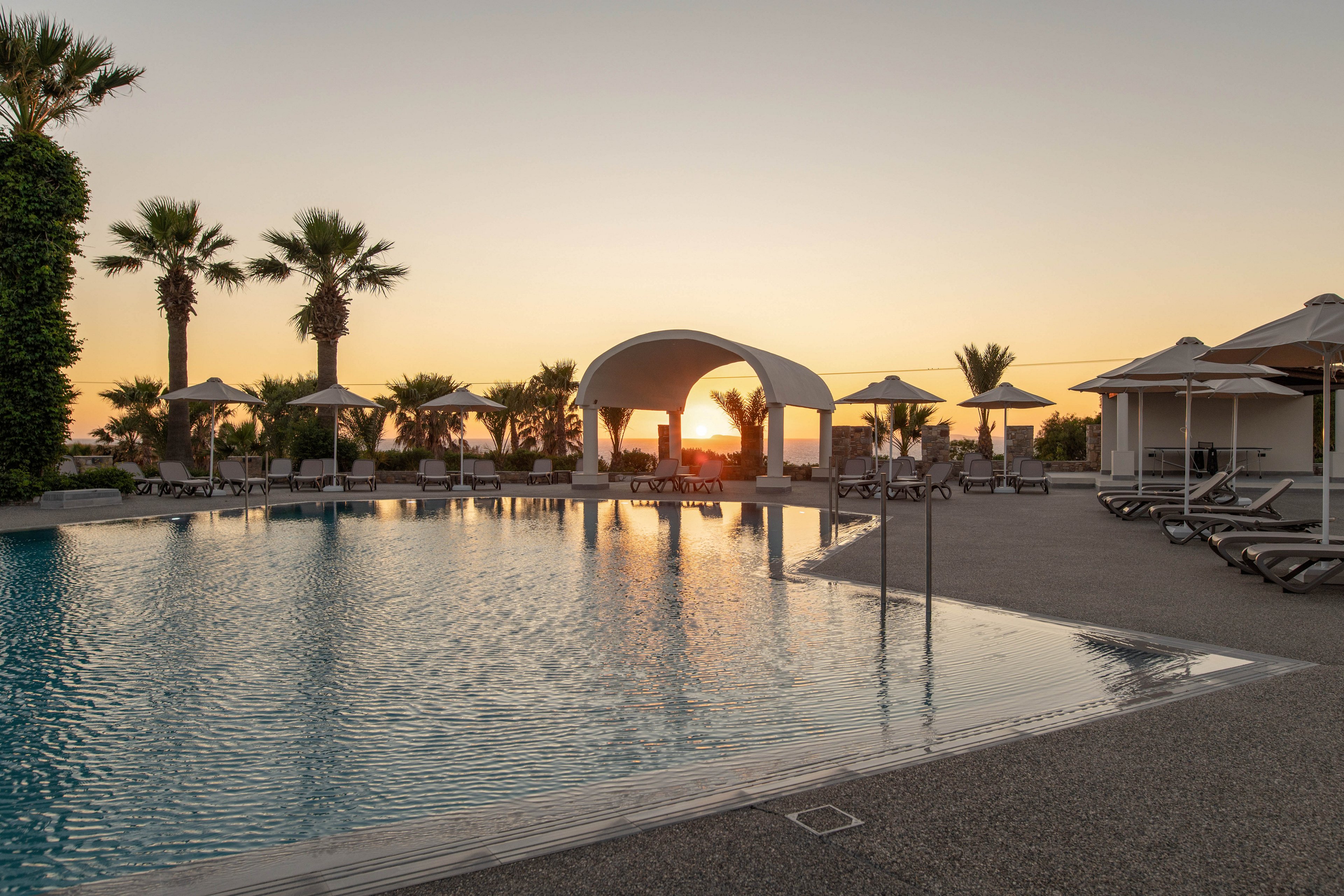 A tranquil resort swimming pool is captured at sunset, with calm water reflecting the warm sky. Palm trees and sun loungers surround the pool, creating a relaxing vacation atmosphere. A white arched pavilion stands near the water, framed by parasols and seating areas. The soft golden light enhances the serene and luxurious feel of the outdoor setting.