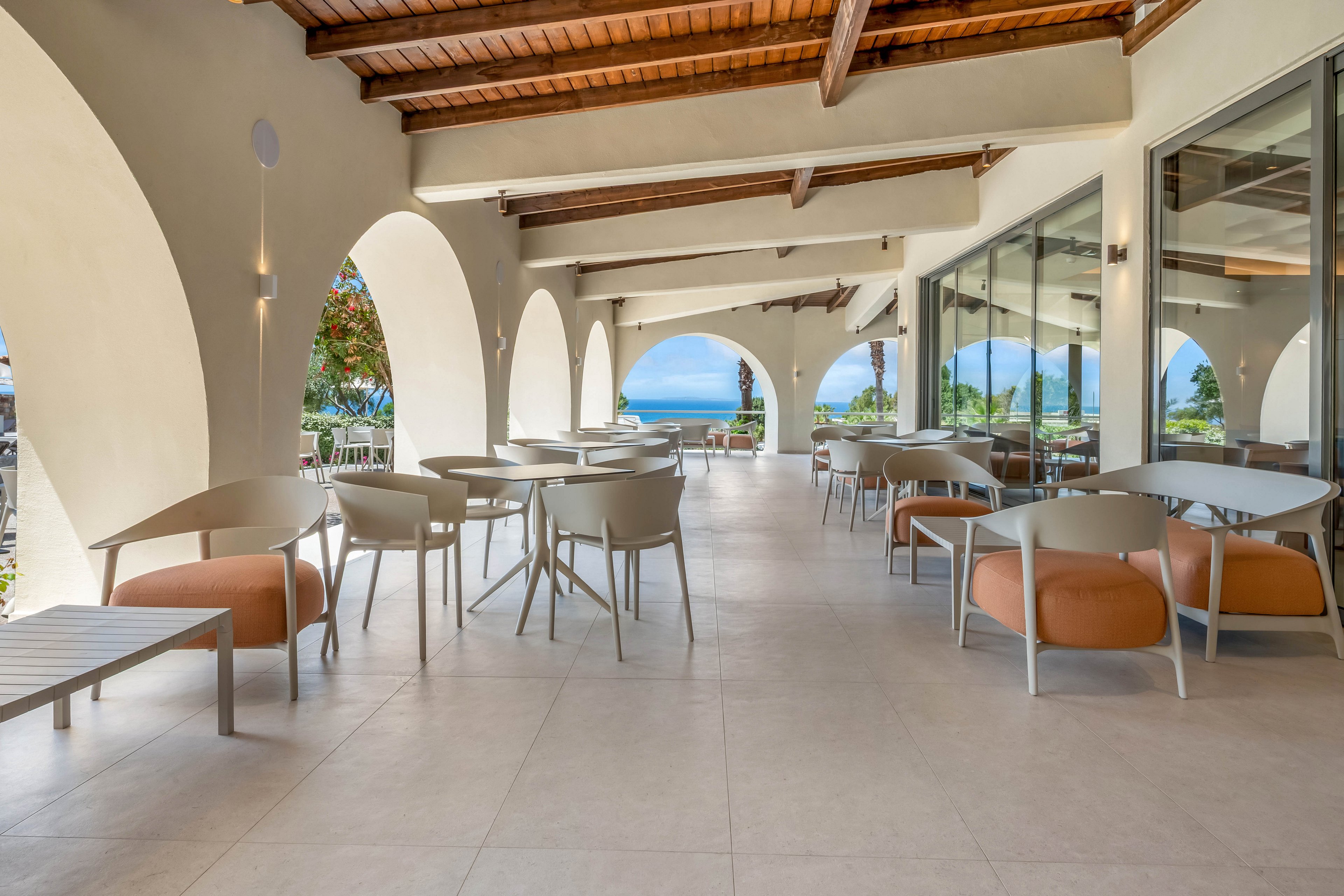 Spacious open-air terrace with contemporary tables and chairs overlooks a distant blue sea. Large white arches frame the view, while a wooden ceiling adds warmth to the bright space. Neutral-toned flooring and minimalist furniture create a clean, modern aesthetic. The setting appears calm and inviting, ideal for outdoor dining or relaxation in a coastal resort.