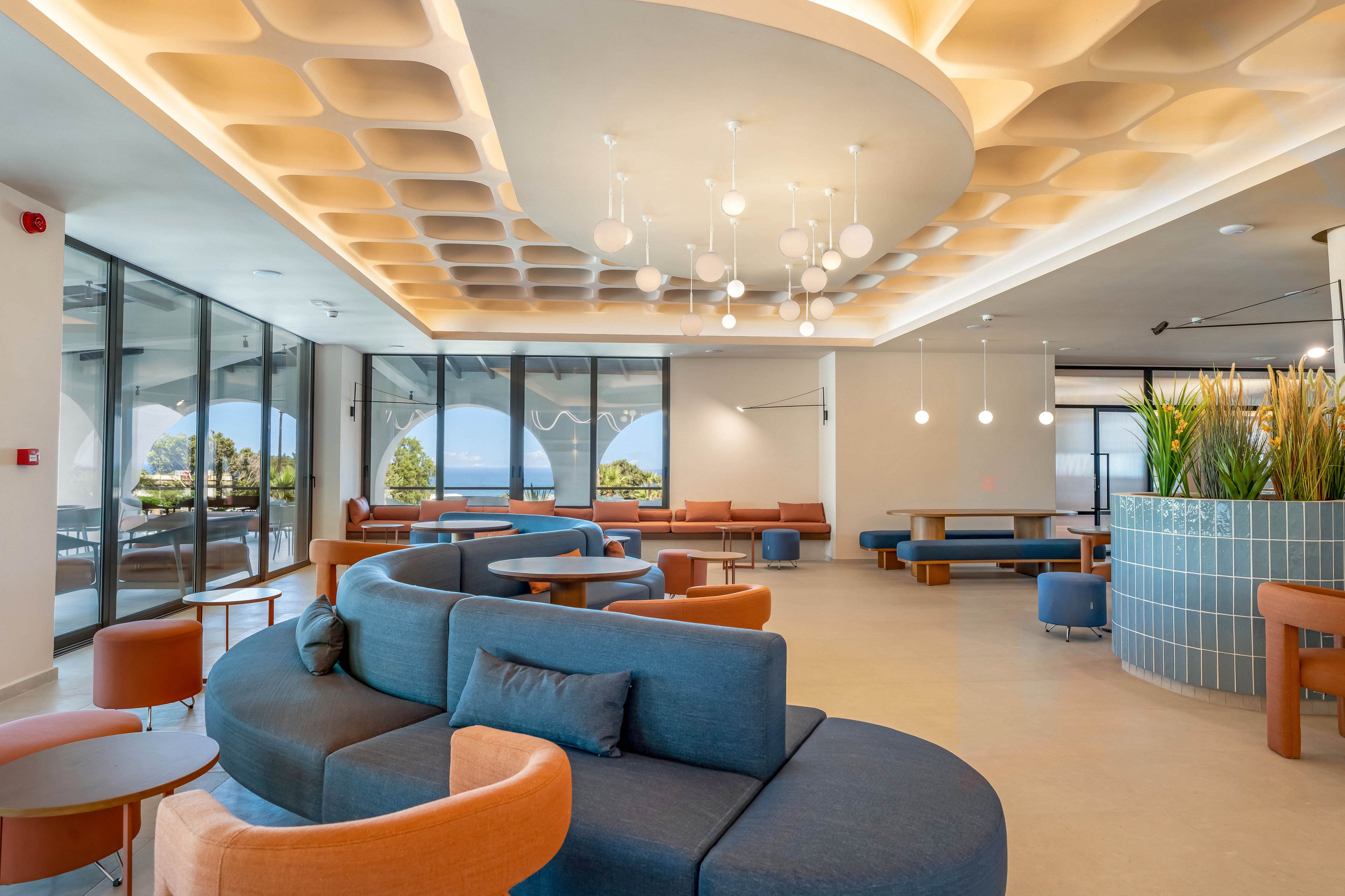 A spacious contemporary lounge features curved modular sofas in blue and orange tones. Large floor-to-ceiling windows open to a coastal view with trees and water outside. The ceiling has a sculpted geometric pattern with recessed lighting and hanging pendant lights. Planters with tall greenery and minimalist side tables add warmth to the clean, modern interior.
