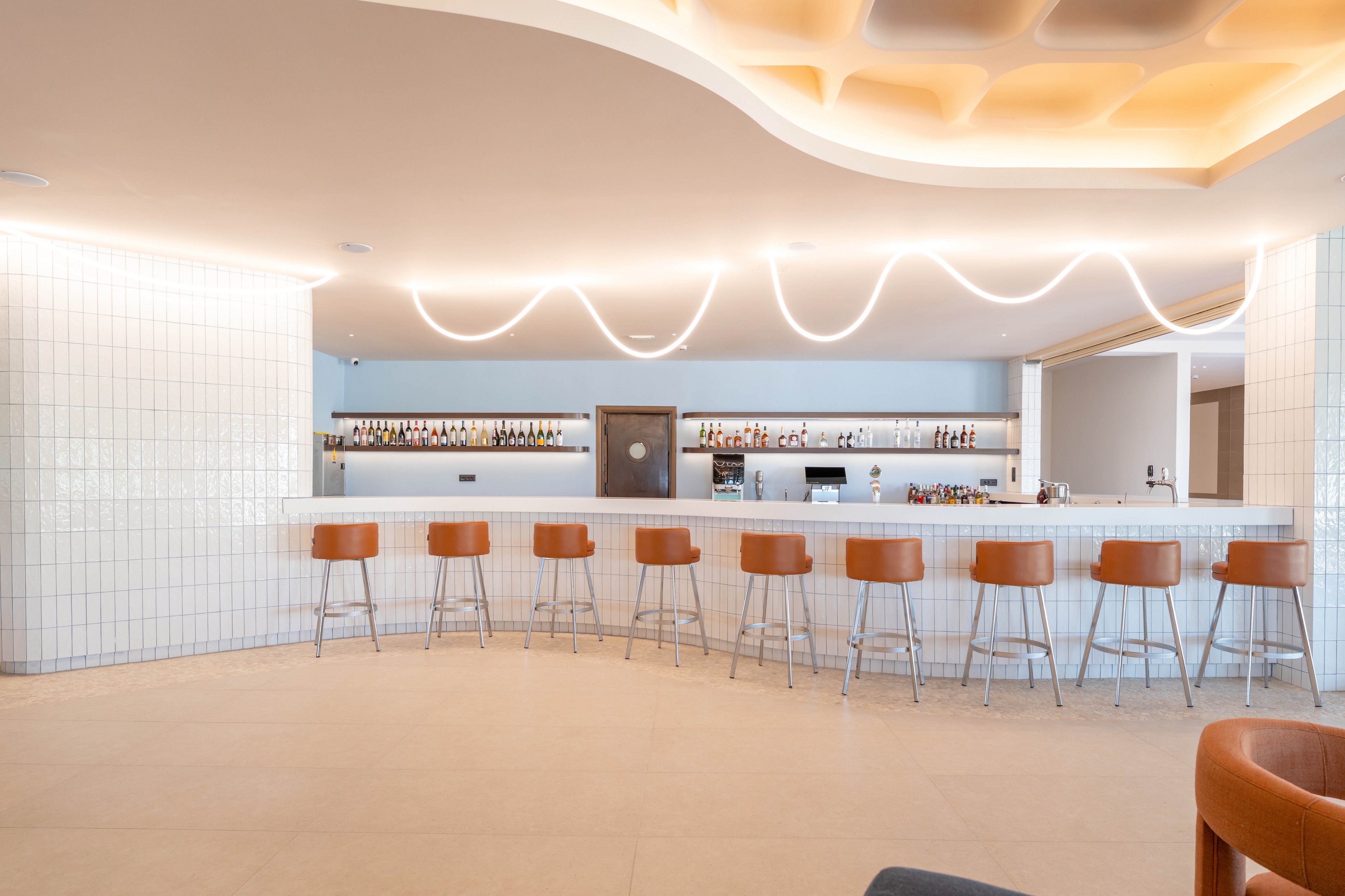 A bright, minimalist bar interior features a long white counter lined with tan bar stools. The space is empty of people, emphasizing clean lines and a calm, orderly atmosphere. Bottles and glassware are neatly arranged on backlit shelves behind the bar. Soft, curved ceiling lights and tiled walls contribute to a contemporary, upscale design aesthetic.
