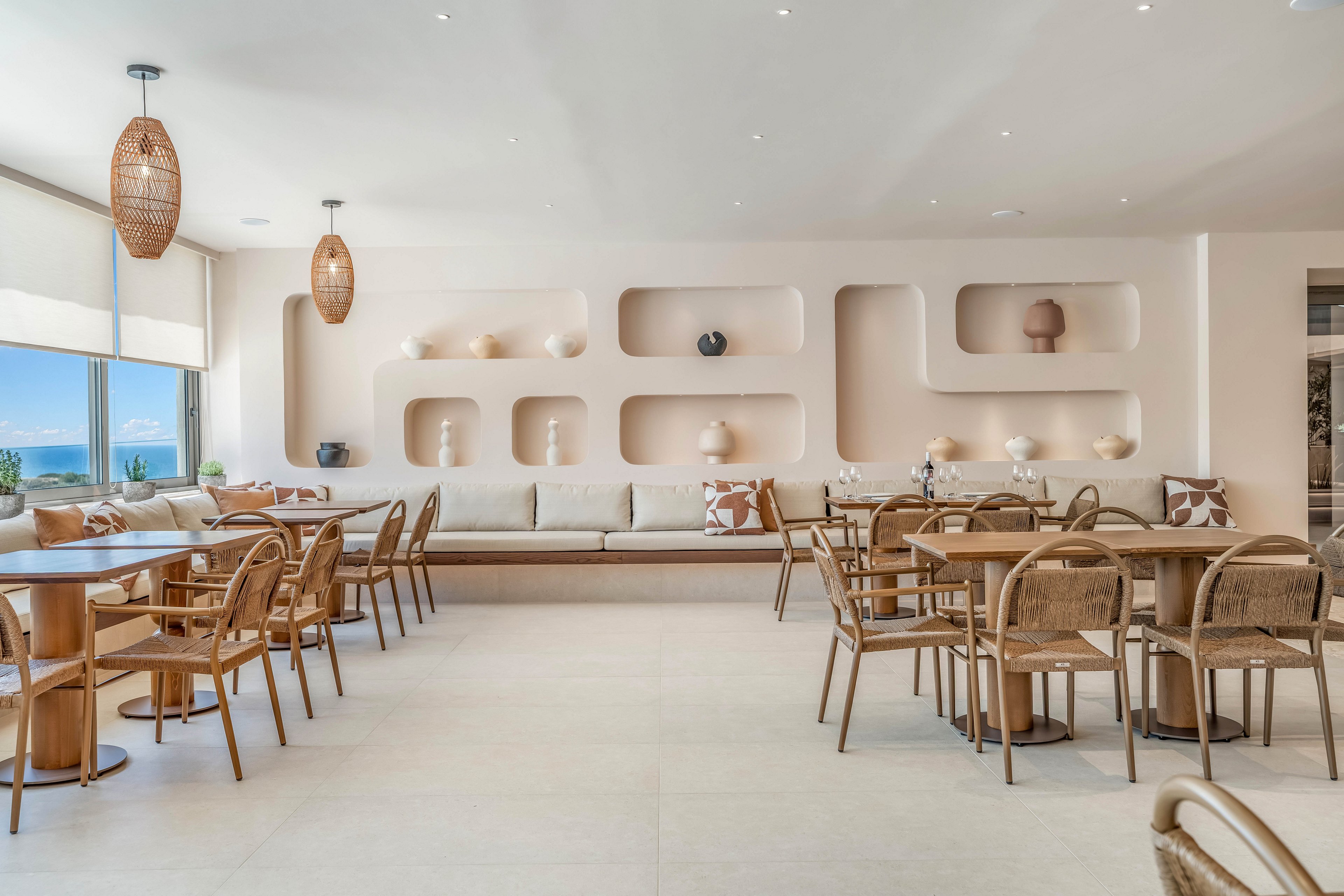 A bright, modern cafe interior features light wood tables and woven chairs arranged neatly across a spacious room. Built-in wall niches display simple decorative vases and pottery, complementing the long upholstered bench along the back wall. Large windows on the left let in natural light and offer a glimpse of the sea and blue sky outside. The neutral color palette and soft lighting create a calm, relaxed atmosphere suitable for dining or casual meetings.