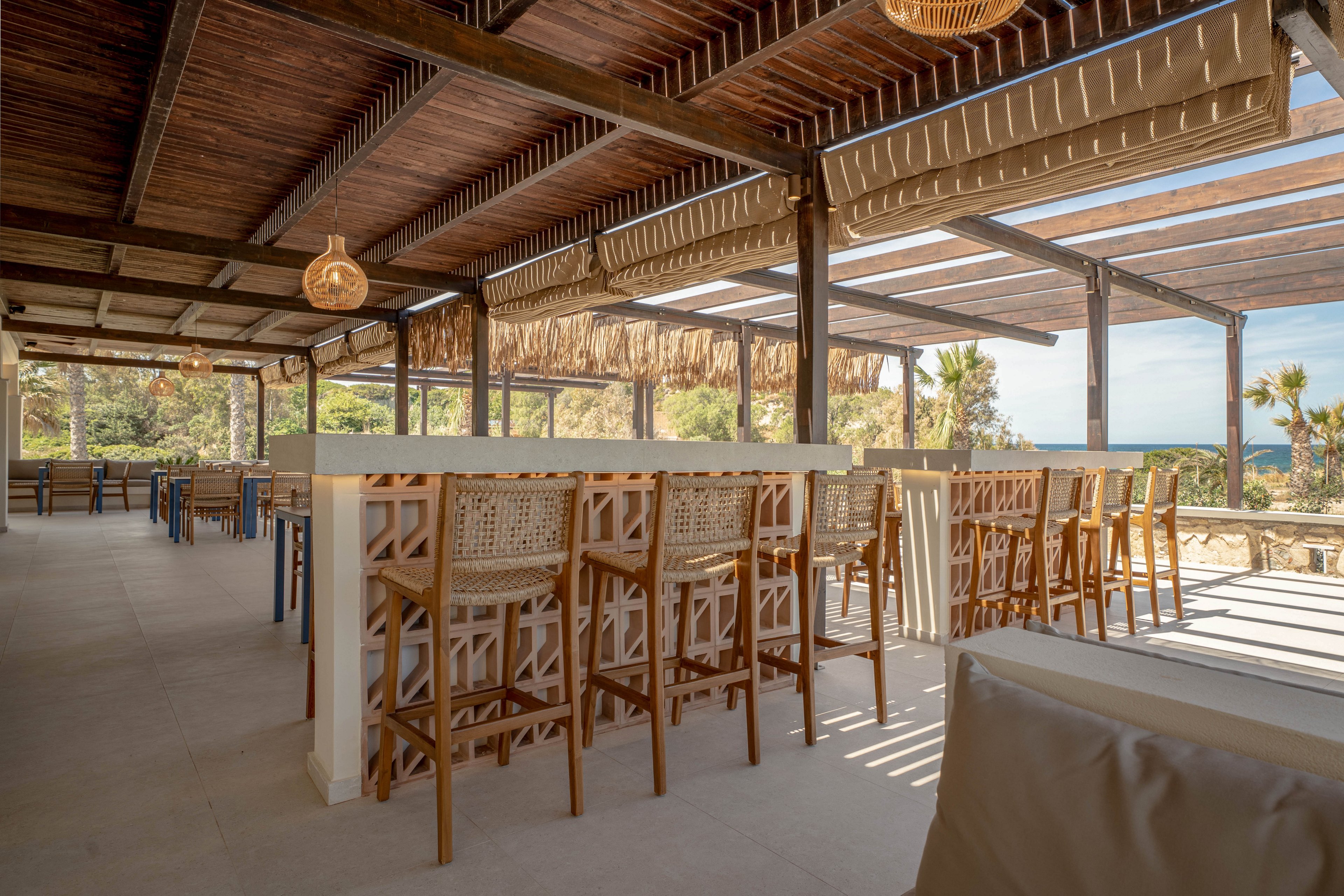 An open-air bar with high wooden tables and woven bar stools sits under a shaded pergola. The space features natural materials, including a wooden ceiling, wicker lamps, and neutral-toned flooring. Large openings reveal palm trees and a view toward the sea in the background. The overall atmosphere is relaxed and inviting, ideal for coastal dining or drinks.
