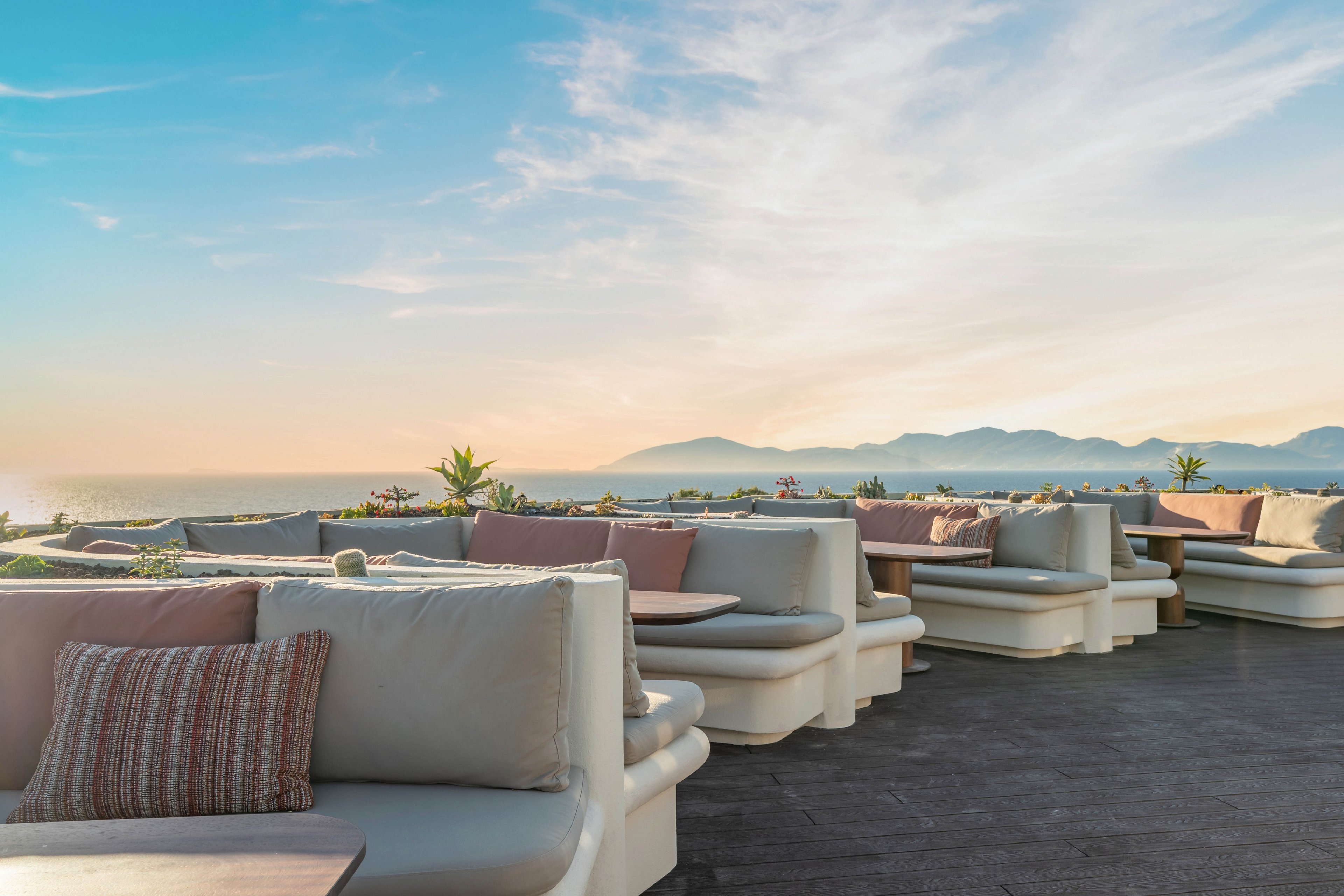 An open-air rooftop lounge features modern cushioned sofas arranged in cozy seating areas. Soft neutral tones with pastel pillows create a relaxed and inviting atmosphere. The terrace overlooks a calm sea with distant mountains under a soft evening sky. Warm natural light enhances the tranquil, resort-like setting.