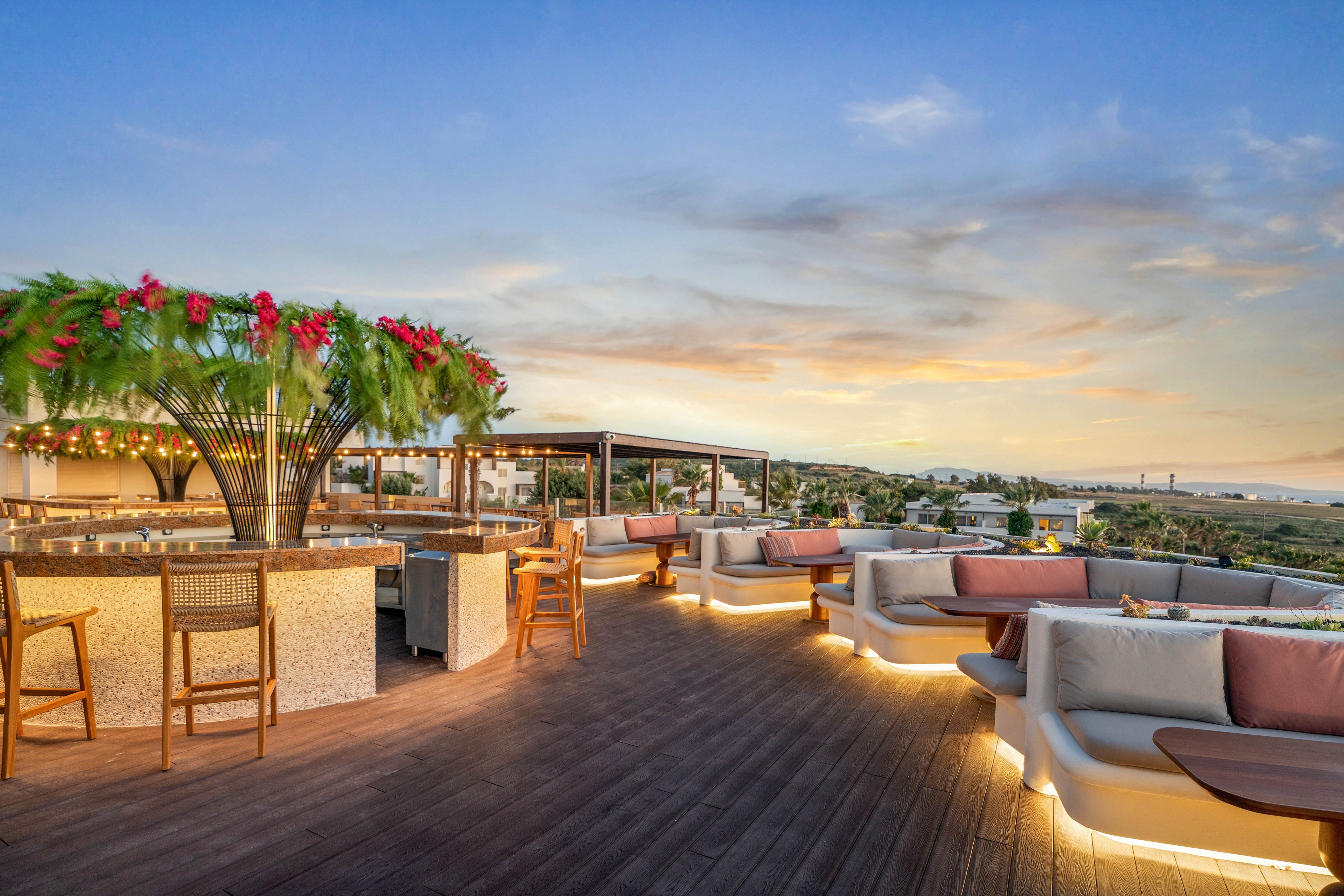 An elegant rooftop lounge bar features cushioned seating areas and a central bar adorned with lush greenery and pink flowers. Warm accent lighting traces the edges of the wooden deck, creating an inviting atmosphere as day turns to dusk. The open-air setting overlooks a distant landscape under a soft, colorful evening sky. The scene suggests a relaxed, upscale environment ideal for socializing and enjoying views.