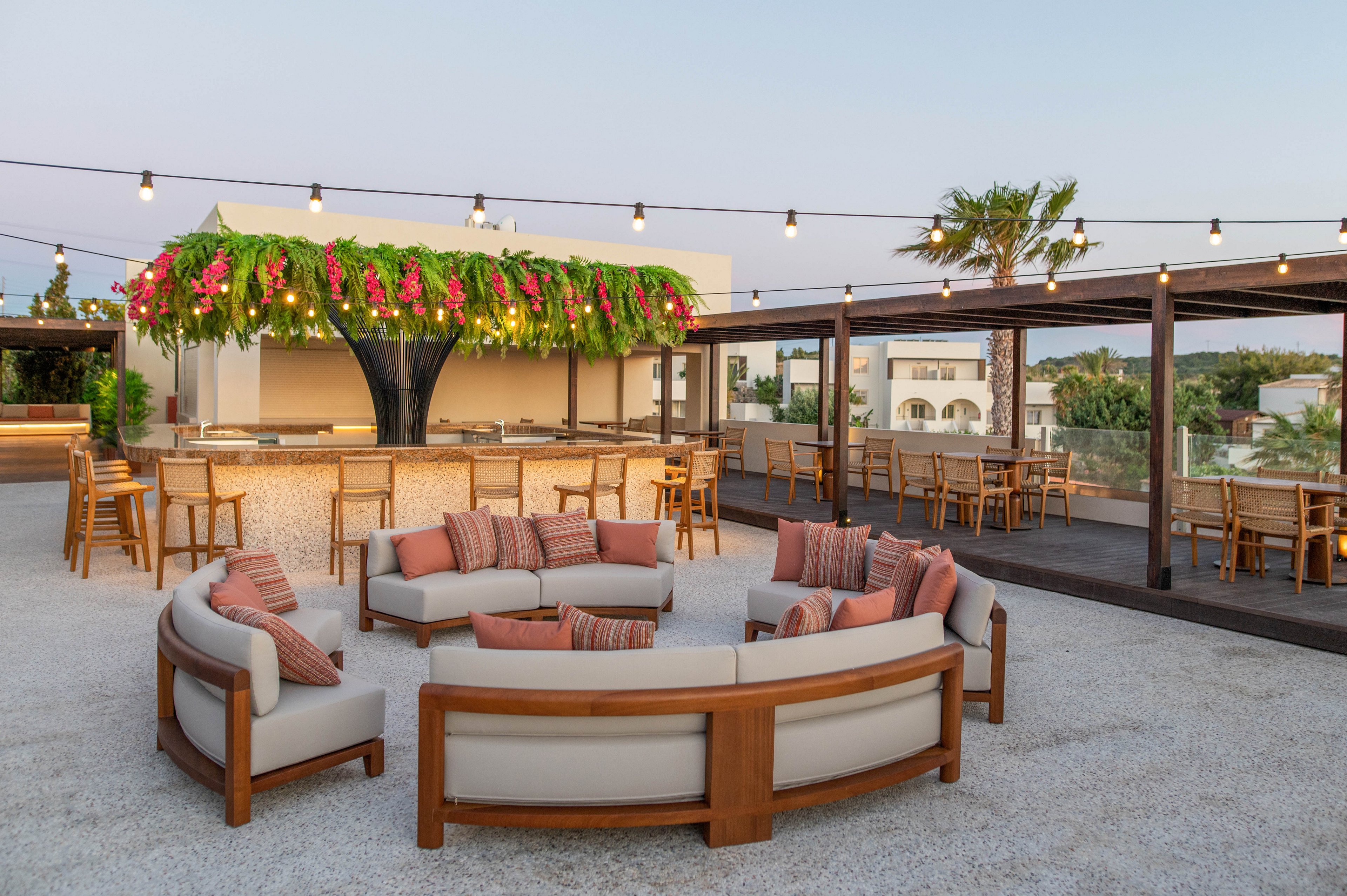 An open-air rooftop lounge features a circular arrangement of cushioned wooden sofas around a central space. In the background, a bar with high stools is shaded by a large canopy covered in lush greenery and pink flowers. String lights are suspended overhead, adding ambiance as evening approaches. Additional bar seating and distant buildings are visible under a clear sky, creating a relaxed, upscale outdoor setting.