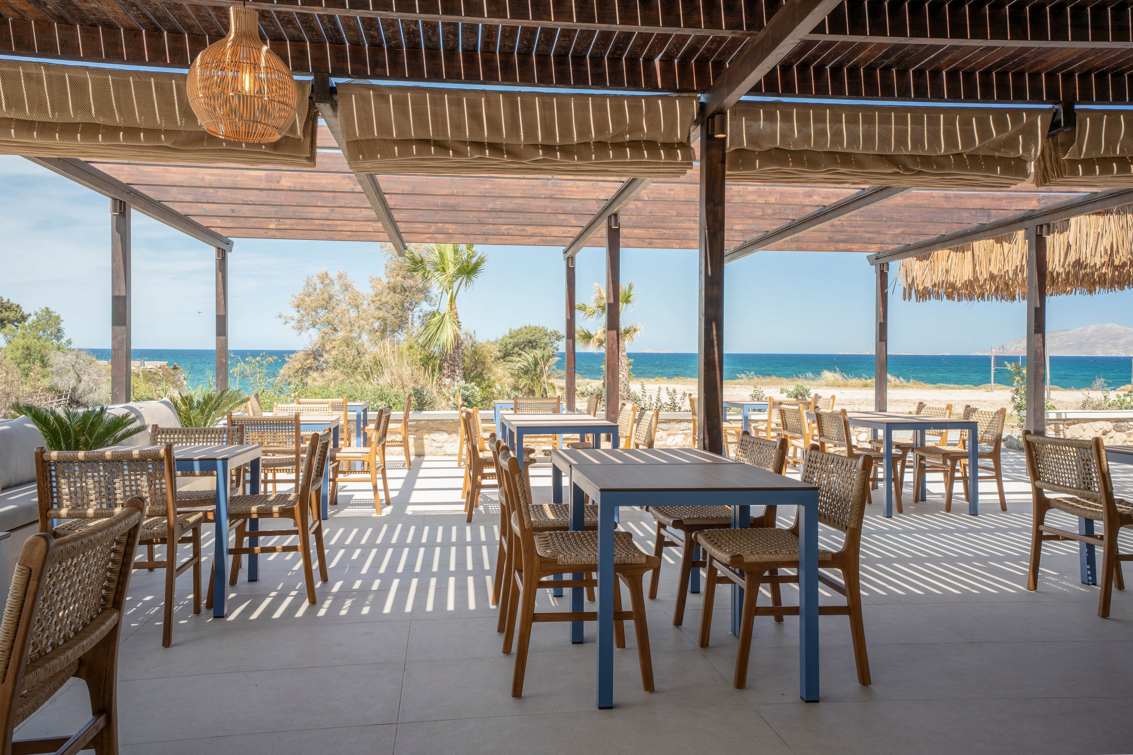 An open-air café with wooden tables and woven chairs overlooks a sandy beach and blue sea. The seating area is shaded by a slatted pergola that casts striped shadows on the floor. Palm trees and coastal vegetation frame the view toward the water and distant horizon. The scene feels calm and inviting, suggesting a relaxed seaside dining experience.