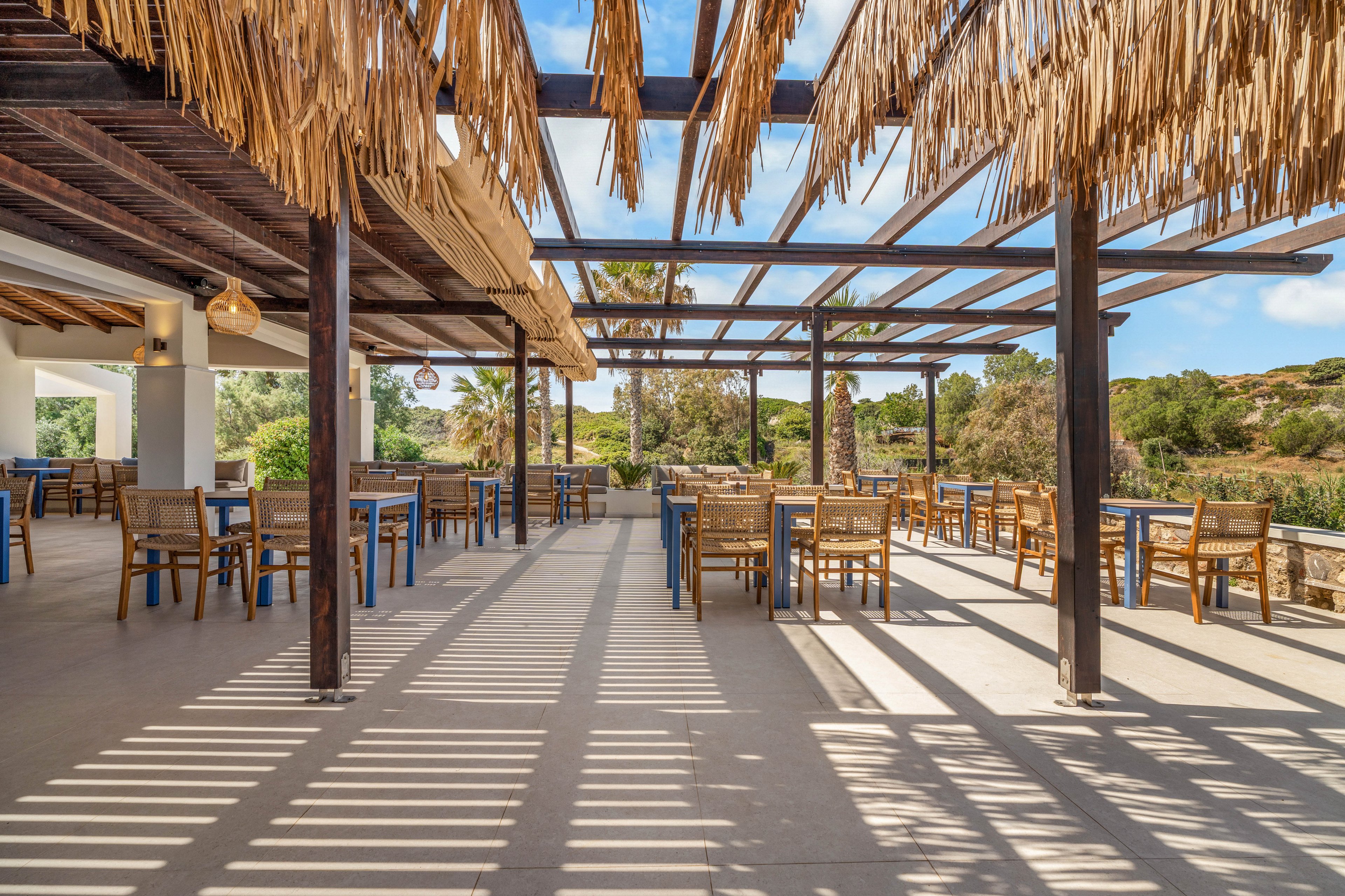 An open-air restaurant terrace features wooden tables and wicker chairs neatly arranged under a pergola. Sunlight filters through the slatted roof, casting strong linear shadows across the floor. The space overlooks lush greenery and trees under a bright blue sky with scattered clouds. The scene feels calm and inviting, ideal for relaxed outdoor dining in warm weather.