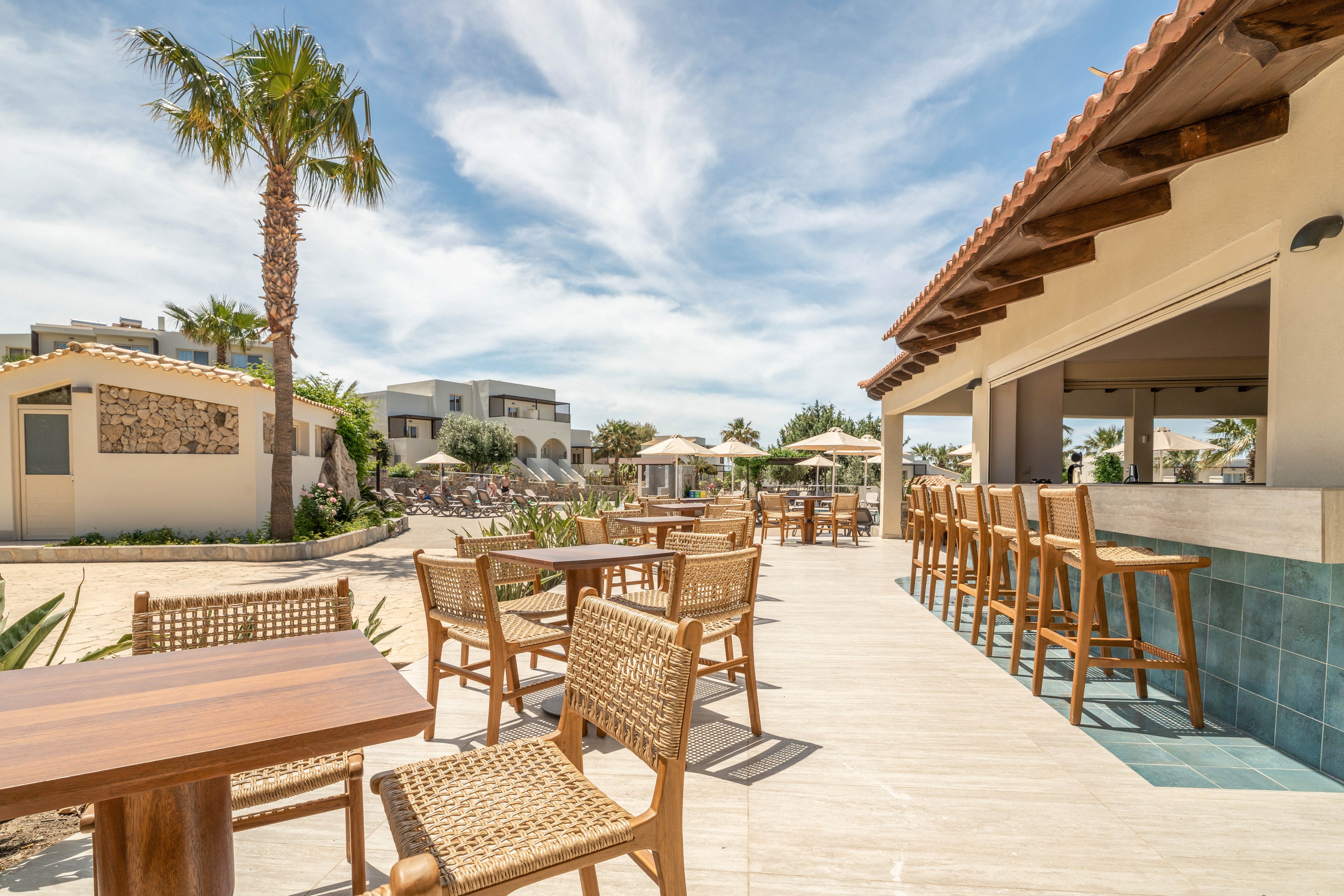 Outdoor wooden tables and wicker chairs sit on a spacious terrace at a beach resort bar. High stools line the bar counter under a tiled roof, facing the open sandy courtyard. Modern low-rise buildings and bungalows with stone accents appear in the background. A tall palm tree and clear blue sky with wispy clouds create a relaxed, vacation atmosphere.