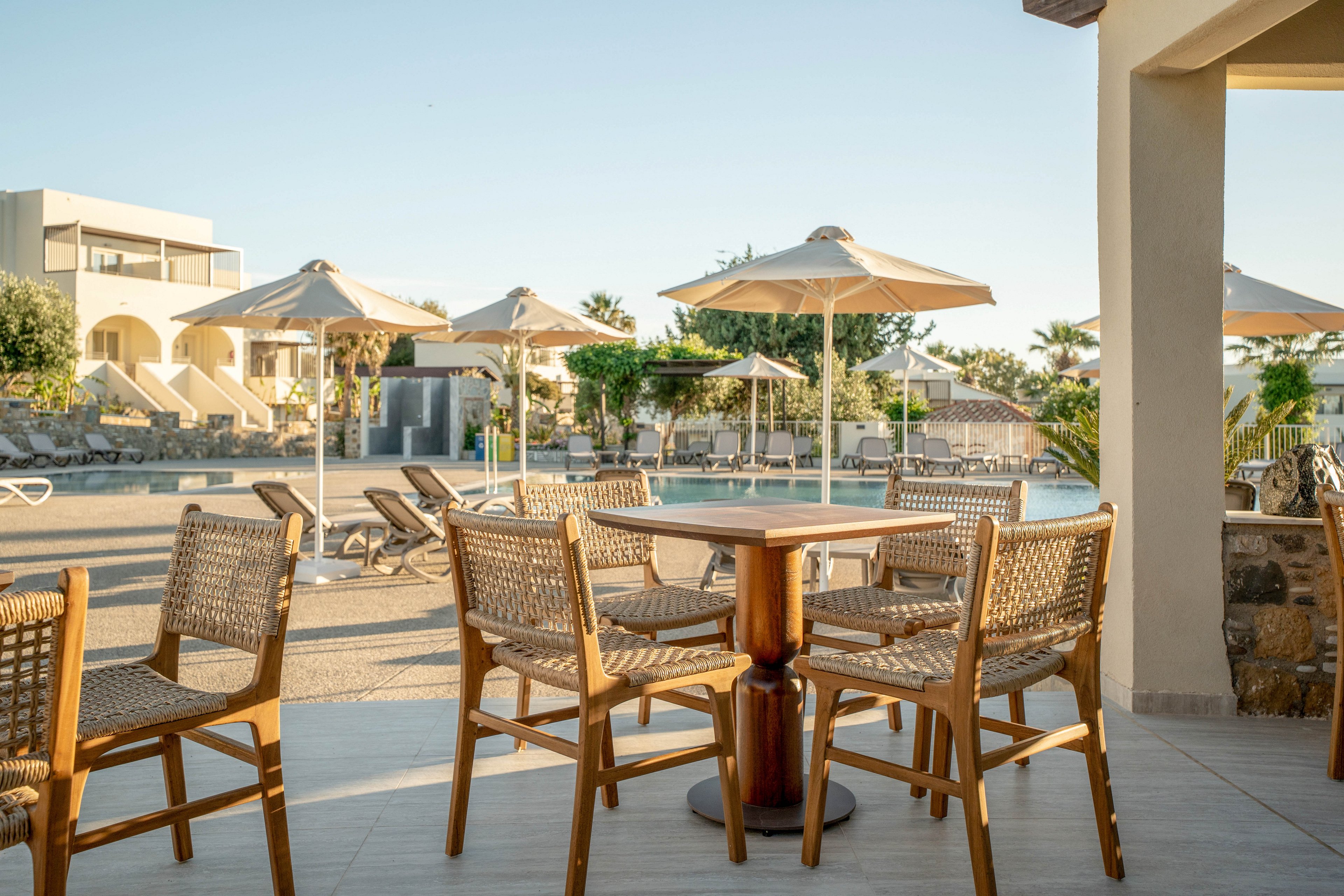 An outdoor resort terrace features a wooden table surrounded by wicker chairs. In the background, a clear swimming pool is lined with sun loungers and large white umbrellas. Modern low-rise buildings and palm trees frame the scene under a clear blue sky. The warm sunlight suggests a calm, relaxing vacation atmosphere.