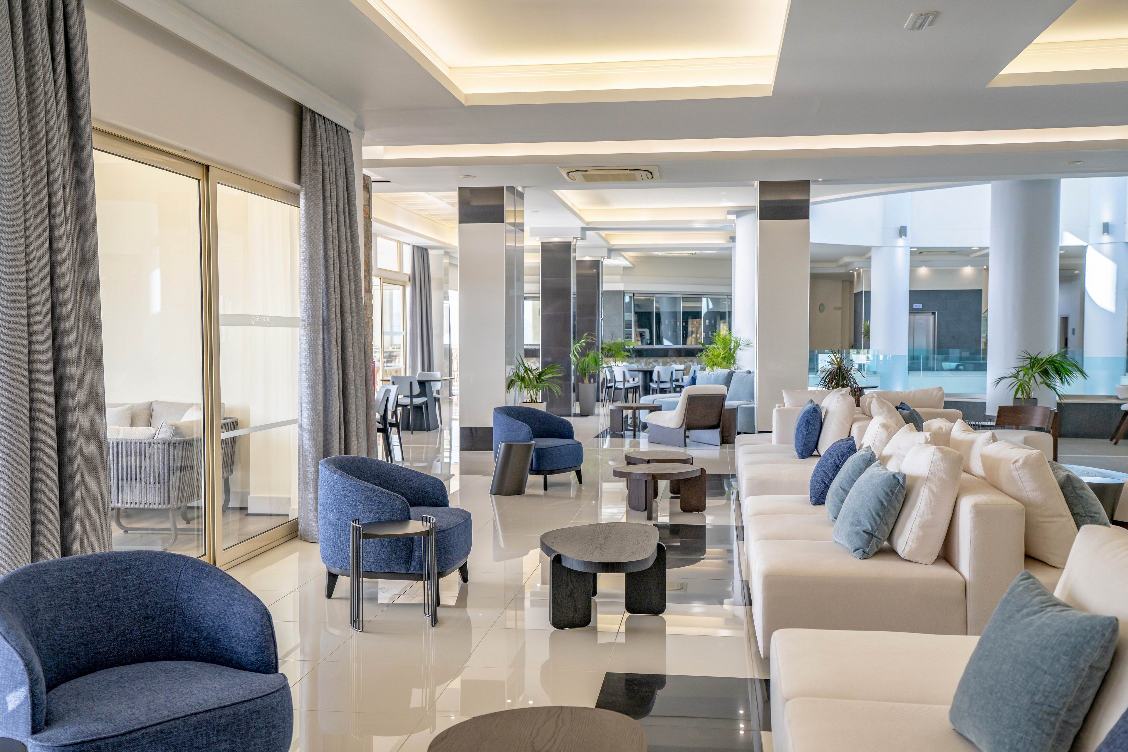 Bright contemporary lounge area featuring cushioned sofas and modern armchairs in neutral and blue tones. The space includes low coffee tables arranged on a glossy tiled floor, with large windows and glass doors providing natural light. Ceiling recess lighting and sleek columns contribute to the clean, minimalist design. Potted plants and distant seating areas add depth and a welcoming atmosphere.