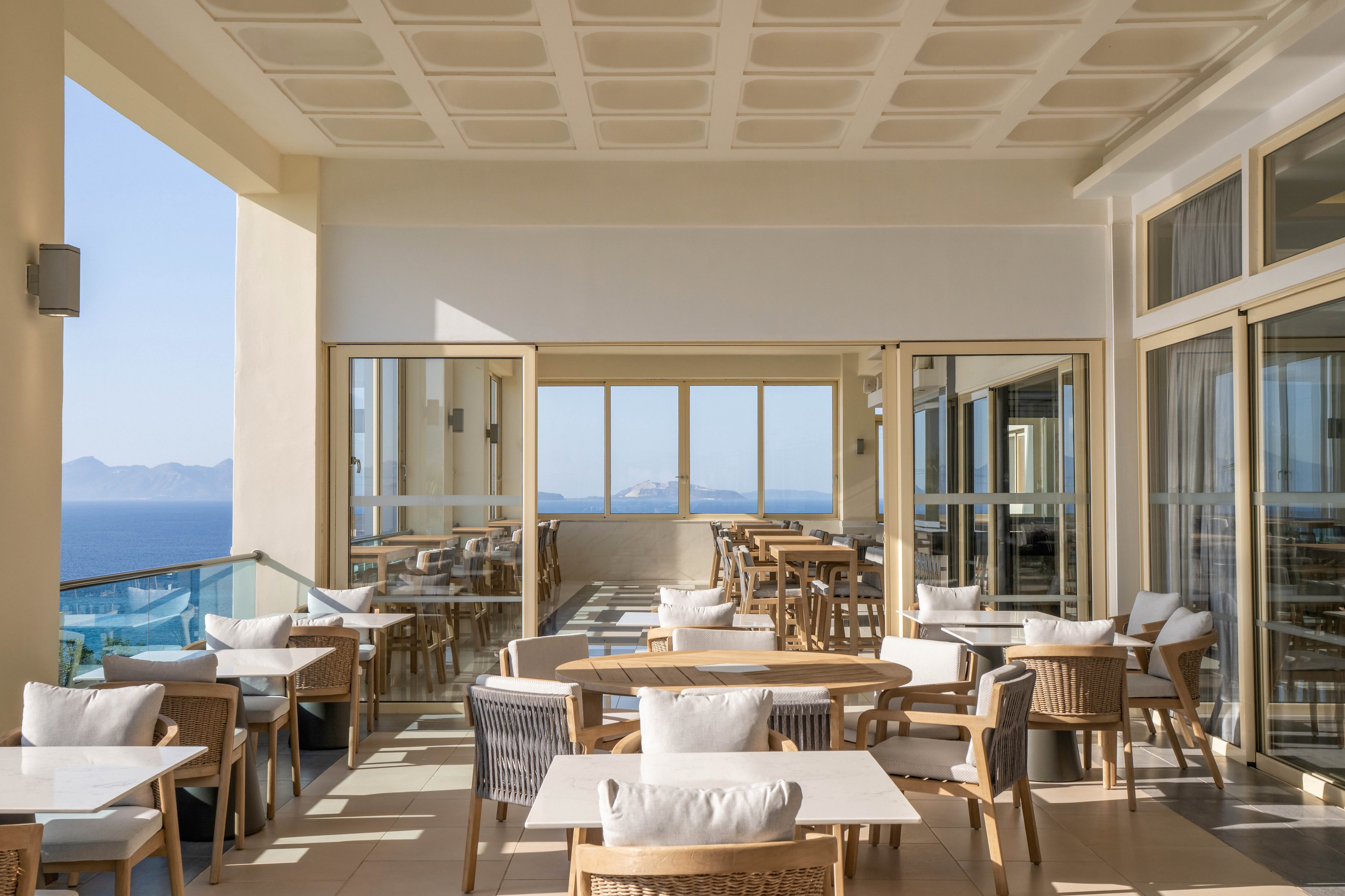 Bright outdoor restaurant terrace overlooking a calm blue sea. Wooden chairs and white tables are neatly arranged, with soft cushions creating a comfortable seating area. Large glass doors and railings frame the expansive coastal view under a structured ceiling. The scene feels peaceful and inviting, ideal for relaxed dining by the water.