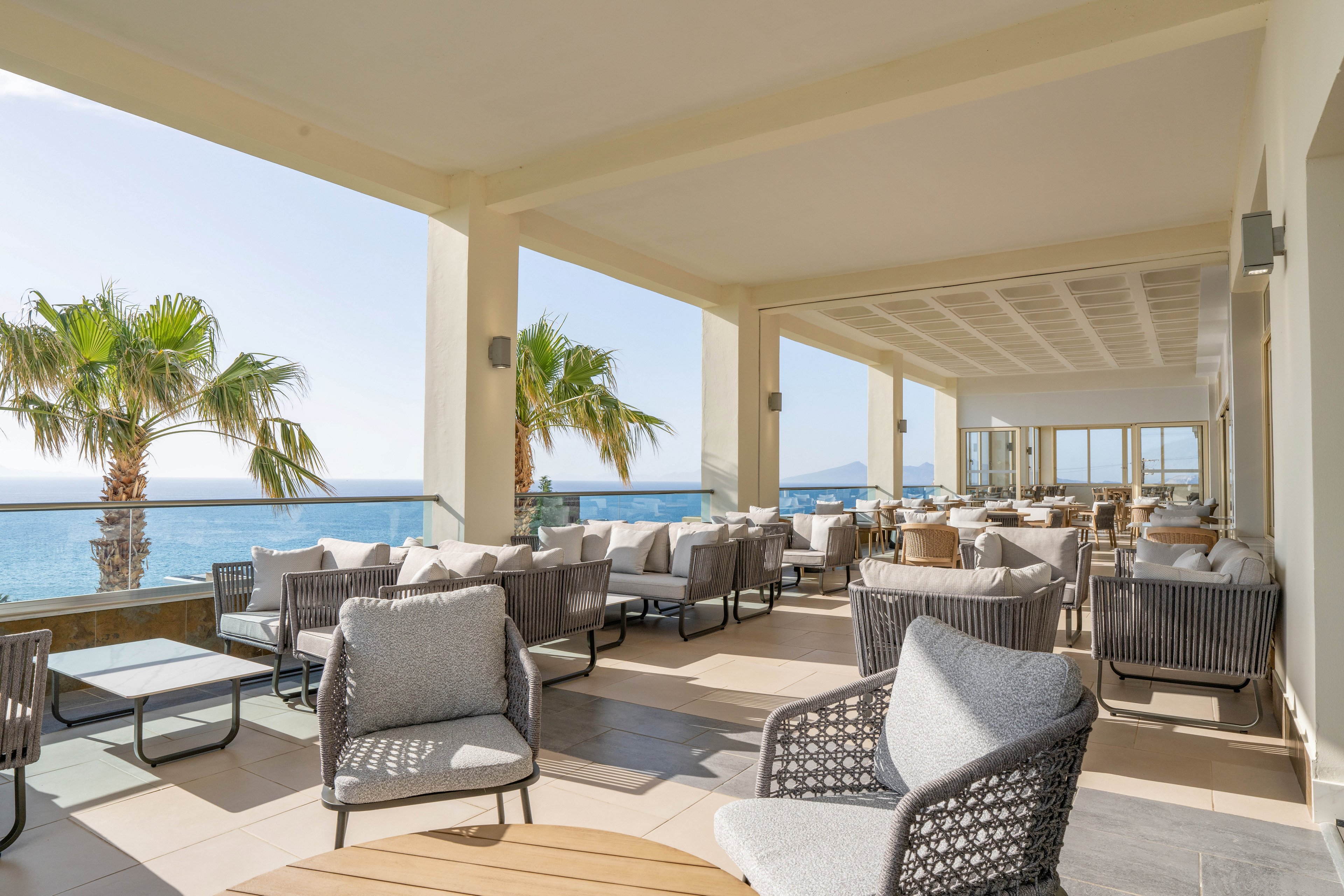 Open-air terrace with contemporary cushioned lounge chairs overlooks a calm blue sea. Palm trees line the edge of the patio, adding a tropical touch to the coastal setting. Neutral-toned furniture and soft textiles create a relaxed, upscale atmosphere under the covered pergola. Sunlight floods the space, highlighting the inviting seating areas and ocean views.