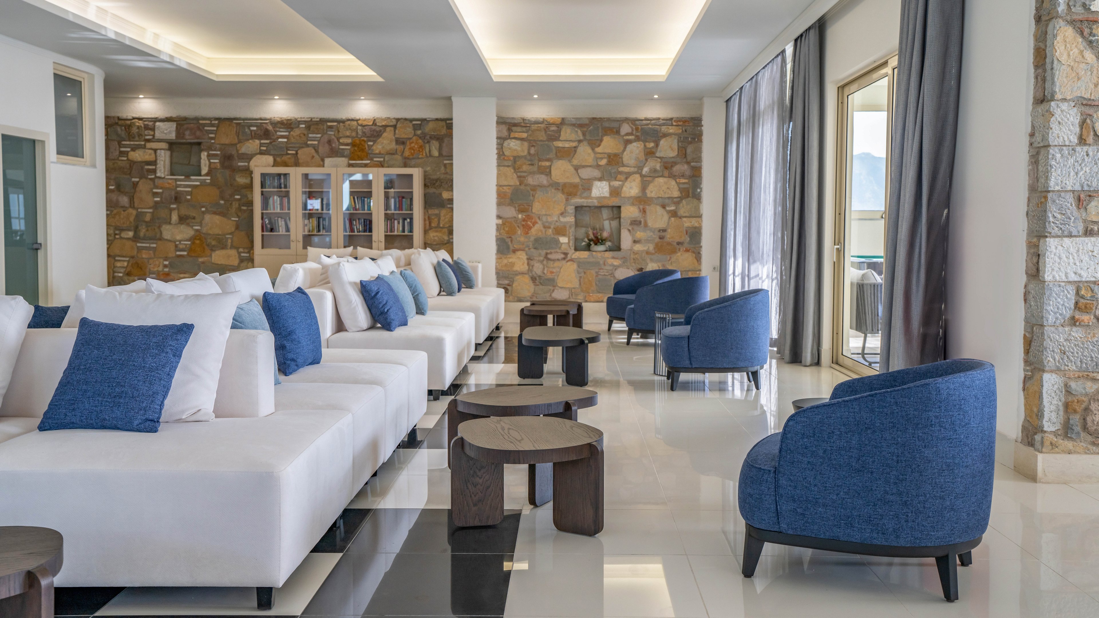 A contemporary lounge area features long white sofas accented with blue and white cushions. Matching blue armchairs and dark wooden side tables are arranged on a glossy white floor. Large windows with sheer curtains let in natural light and offer a view outside, while a stone accent wall adds texture to the space. The overall design feels clean, airy, and inviting, suitable for a hotel or upscale residence.