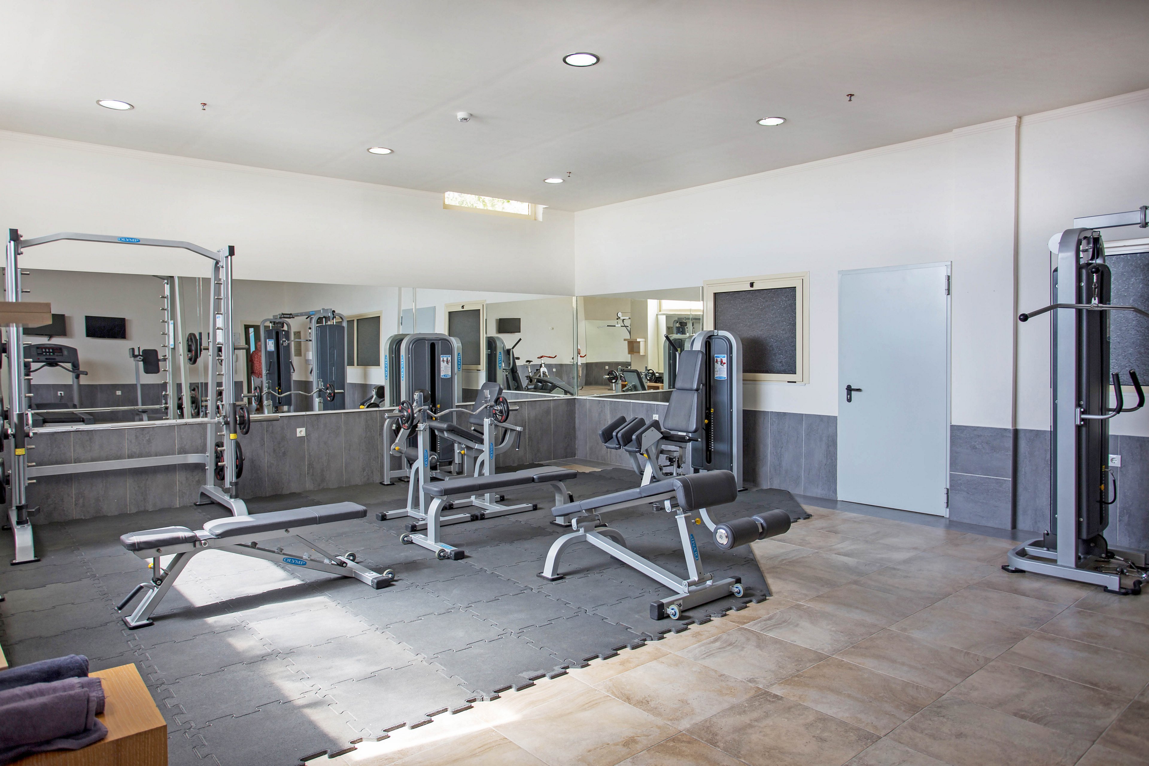 A clean, modern gym room features several strength training machines and weight benches. The space is empty, with no people present, emphasizing the equipment and open workout area. Large mirrors line the wall behind the machines, reflecting the room and adding depth. Neutral-colored flooring and walls create a bright, minimalistic fitness environment.