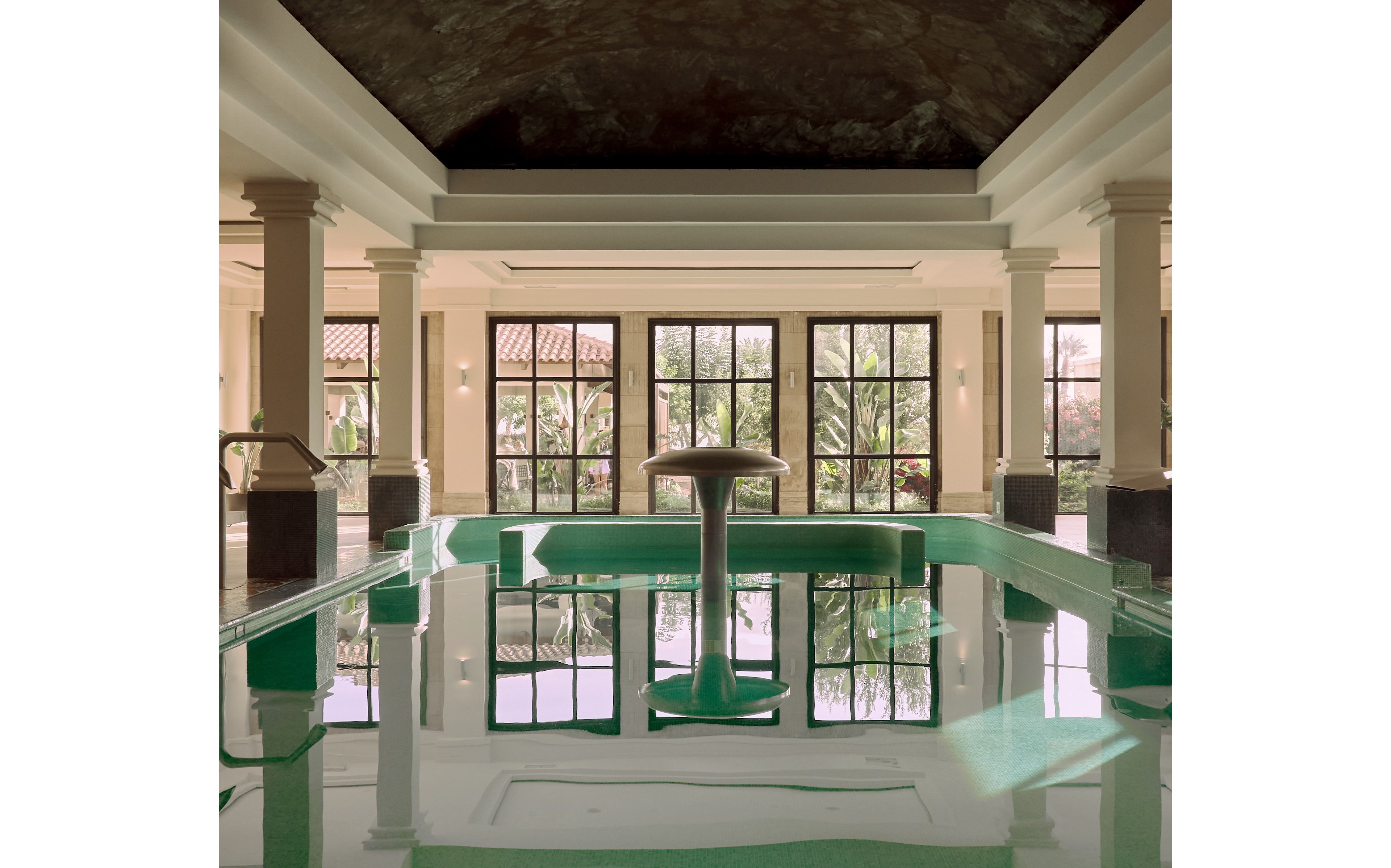 An elegant indoor spa pool features a central mushroom-shaped fountain surrounded by calm, reflective water. Tall columns frame the space, leading the eye toward large grid-style windows at the far end. Sunlight filters through the windows, revealing lush greenery and tiled rooftops outside. The overall atmosphere is serene and luxurious, with soft neutral tones contrasting the green-tinted pool.