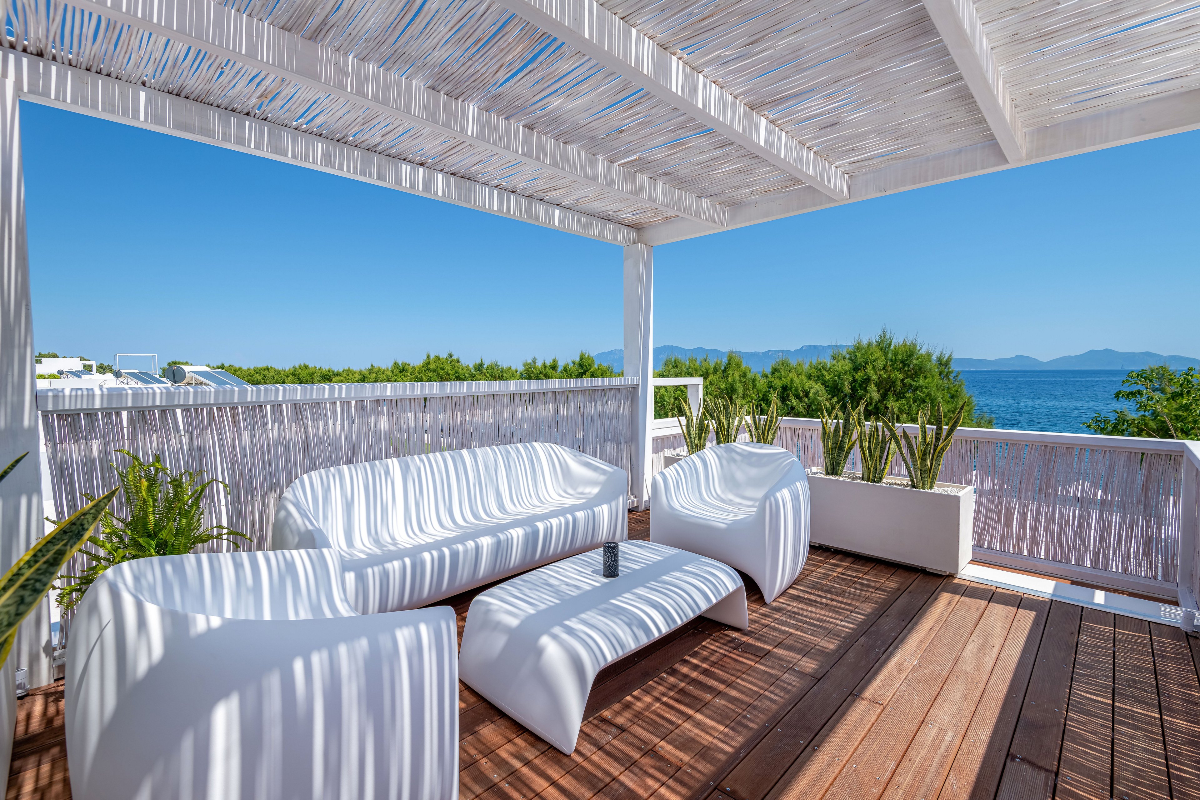 A contemporary outdoor terrace features sculptural white lounge chairs and a matching low table. The space is covered by a white pergola with slatted shading that casts soft linear shadows on the wooden deck. Potted green plants line the railing, framing a clear view of the blue sea and distant mountains. The bright sunlight and vivid blue sky create a fresh, relaxing coastal atmosphere.