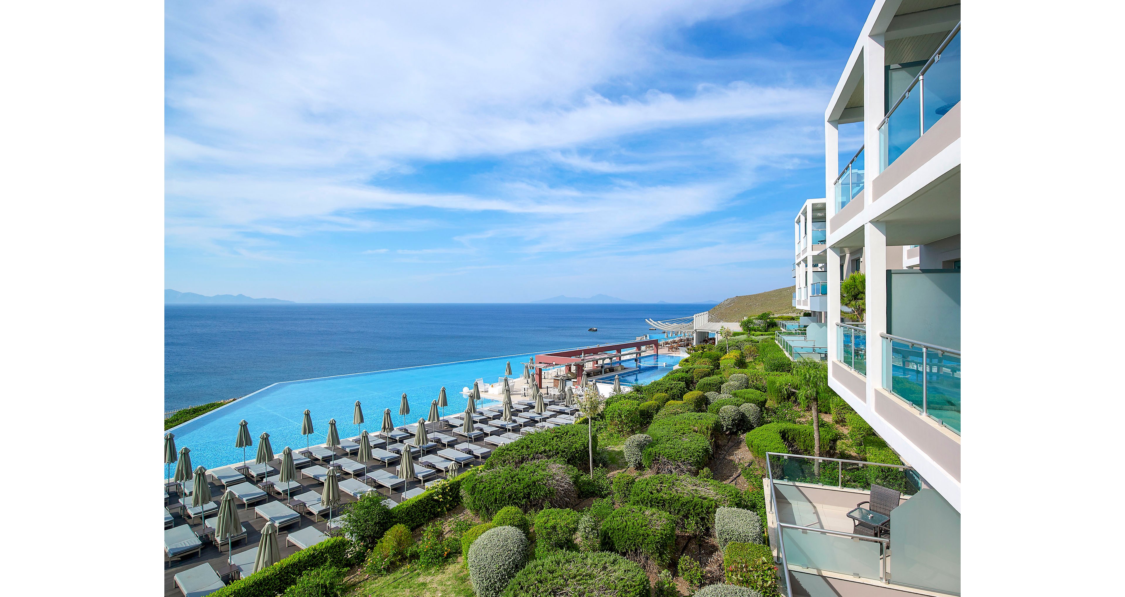 A modern coastal resort overlooks a vast blue sea with an expansive infinity pool in the foreground. Rows of sun loungers and closed umbrellas line the poolside terrace, suggesting a tranquil vacation setting. Contemporary white buildings with glass balconies rise on the right, facing manicured green gardens and shrubs. The clear sky and bright daylight create a serene, relaxing atmosphere ideal for summer holidays.