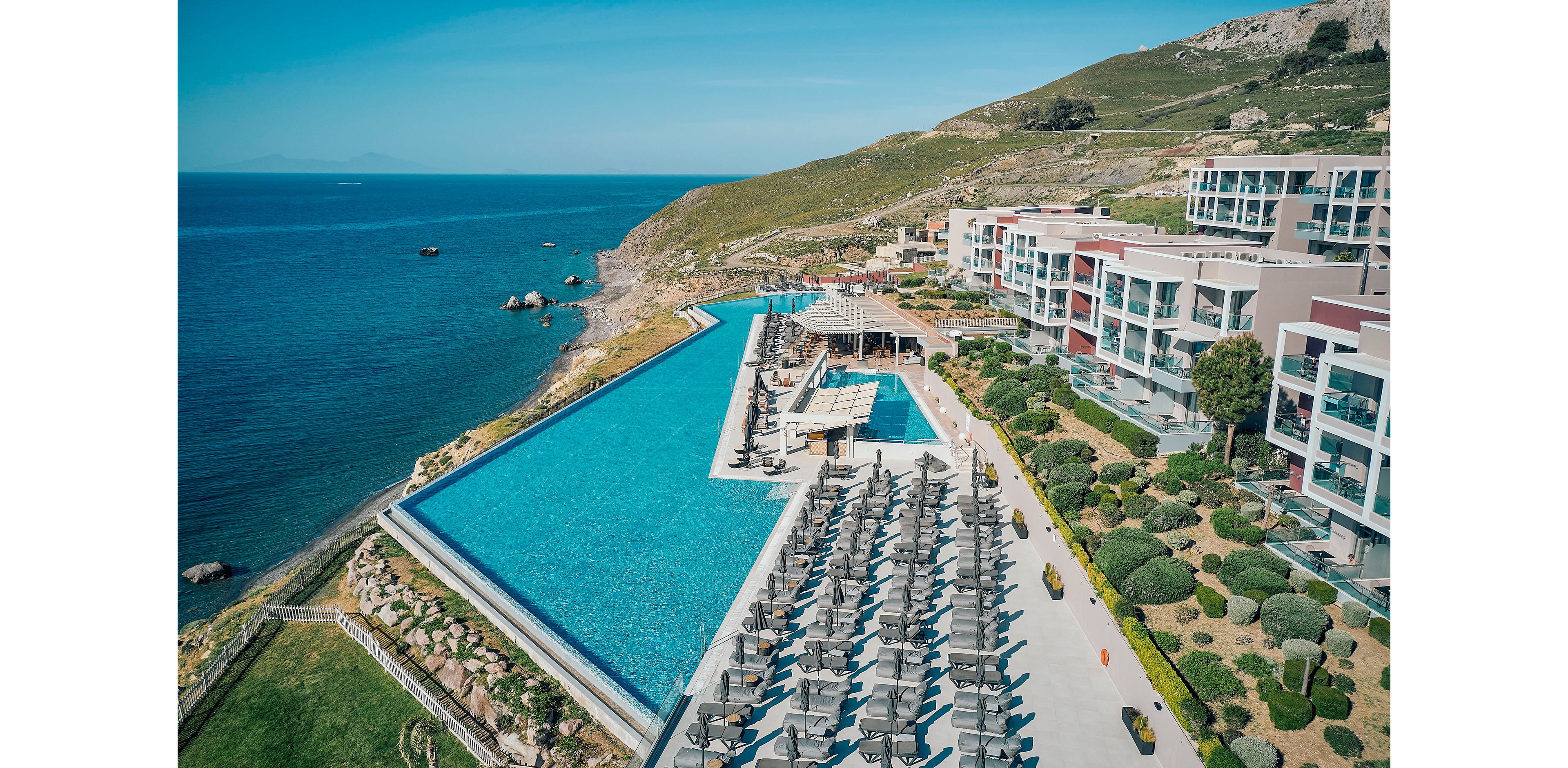 A large luxury resort overlooks a calm blue sea, featuring an expansive infinity pool that stretches along the coastline. Numerous sun loungers and umbrellas are neatly arranged beside the pool, emphasizing a relaxing vacation atmosphere. Modern white and glass buildings rise on the hillside, surrounded by manicured greenery and stone pathways. The scene is captured from above, highlighting the dramatic coastal landscape and clear, sunny weather.