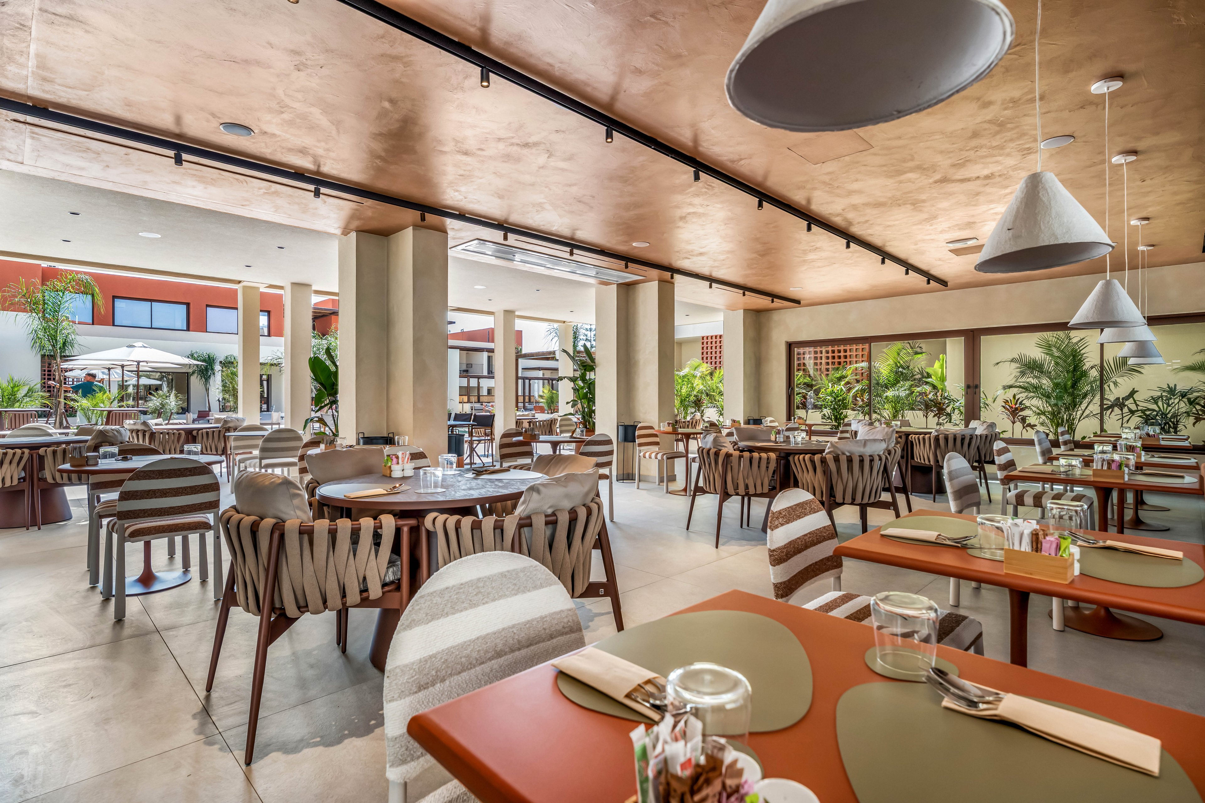 A spacious, modern restaurant dining area is set with neatly arranged tables and chairs. The open-air space features large windows and an outdoor view with lush green plants and palm trees. Neutral tones, wooden accents, and contemporary lighting fixtures create a relaxed yet stylish atmosphere. Table settings with glasses, cutlery, and placemats suggest the restaurant is prepared for guests.