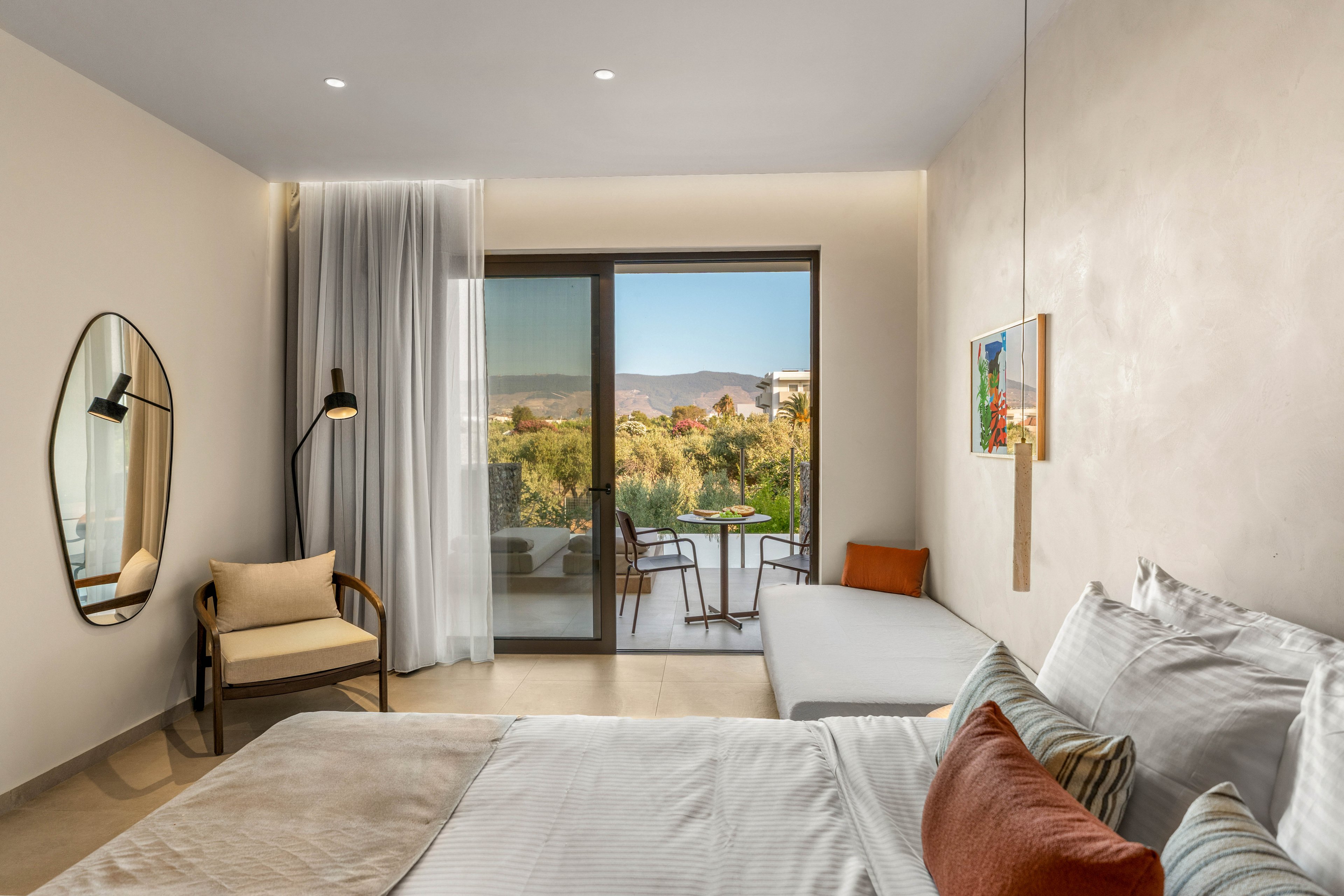 A contemporary hotel room features a neatly made bed with white linens and warm accent pillows. A cozy seating area with a chair and floor lamp sits beside a large glass door leading to a balcony. Outside, a small table and chairs overlook a lush landscape and distant hills under a clear sky. Soft neutral walls, minimalist decor, and natural light create a calm, inviting atmosphere.