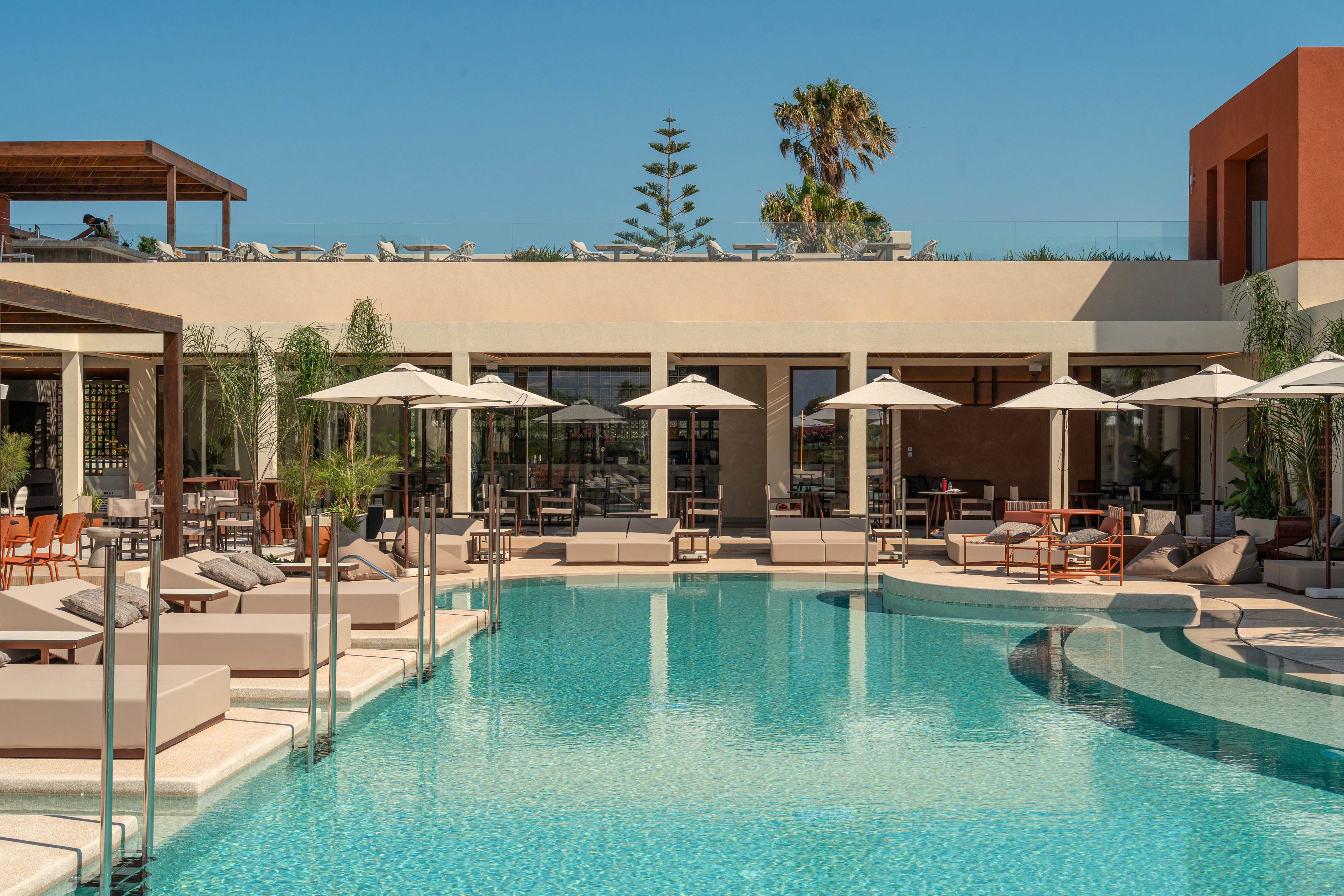 A contemporary resort features a large turquoise swimming pool surrounded by cushioned sun loungers and white umbrellas. The building has clean, minimalist architecture with large glass doors opening onto the pool deck. Palm trees and other greenery add a tropical touch to the outdoor setting. Clear blue skies and bright sunlight create a relaxed vacation atmosphere.