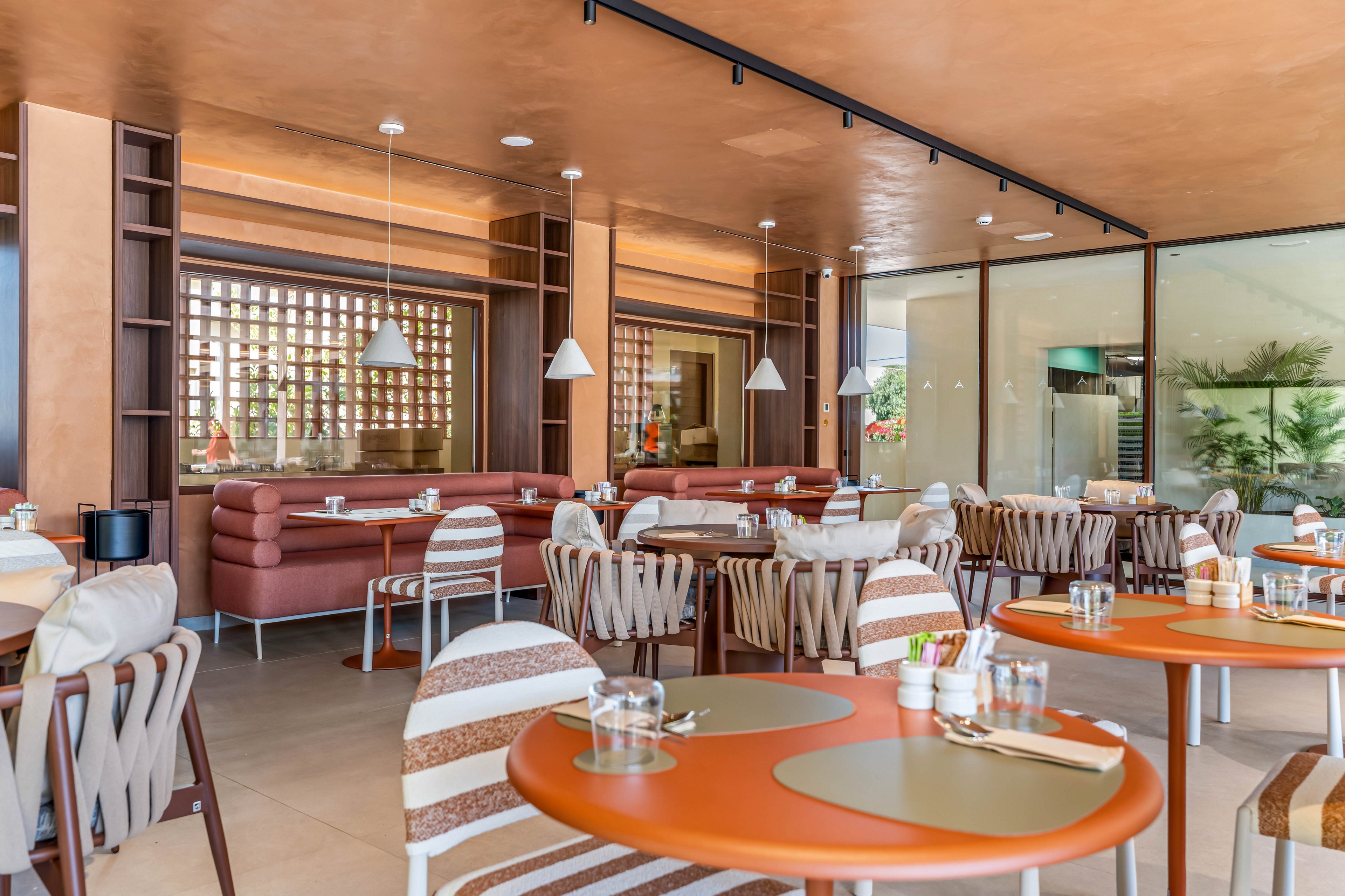 A contemporary restaurant dining area features round orange tables and cushioned chairs neatly arranged in an open layout. Soft pink banquettes line the walls, complemented by wooden shelving and decorative lattice panels. Pendant lights hang from the ceiling, illuminating place settings with glasses and placemats on each table. Large glass windows reveal a bright adjacent space with plants, enhancing the airy and inviting atmosphere.