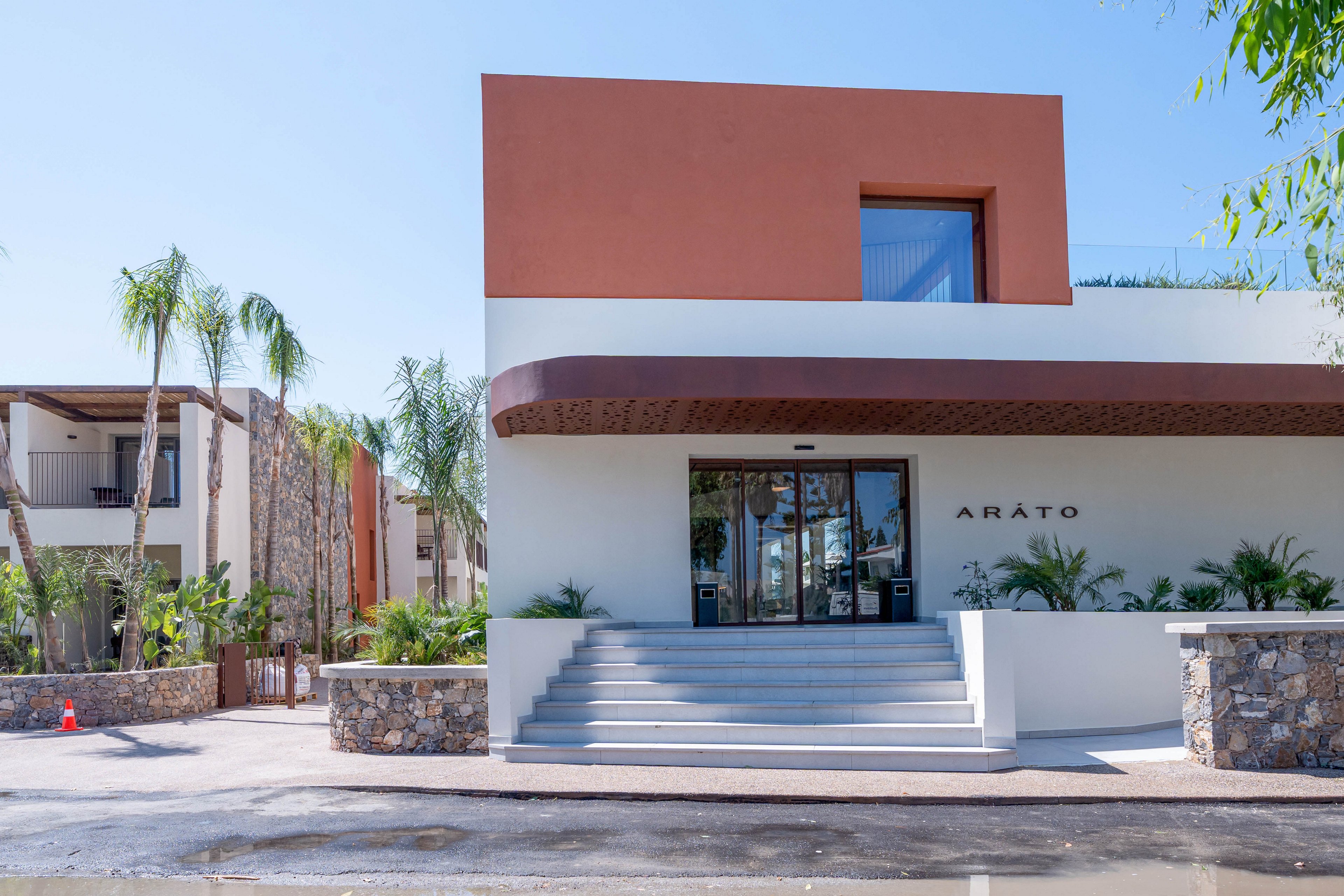 A contemporary building with clean geometric lines serves as the main entrance to a hotel or resort. The facade features white and terracotta-colored sections, with a wide staircase leading up to glass doors. Palm trees and lush greenery surround the structure, adding a tropical atmosphere. Additional buildings and stone walls are visible in the background under a clear blue sky. The word "ARATO" appears on the front wall near the entrance.
