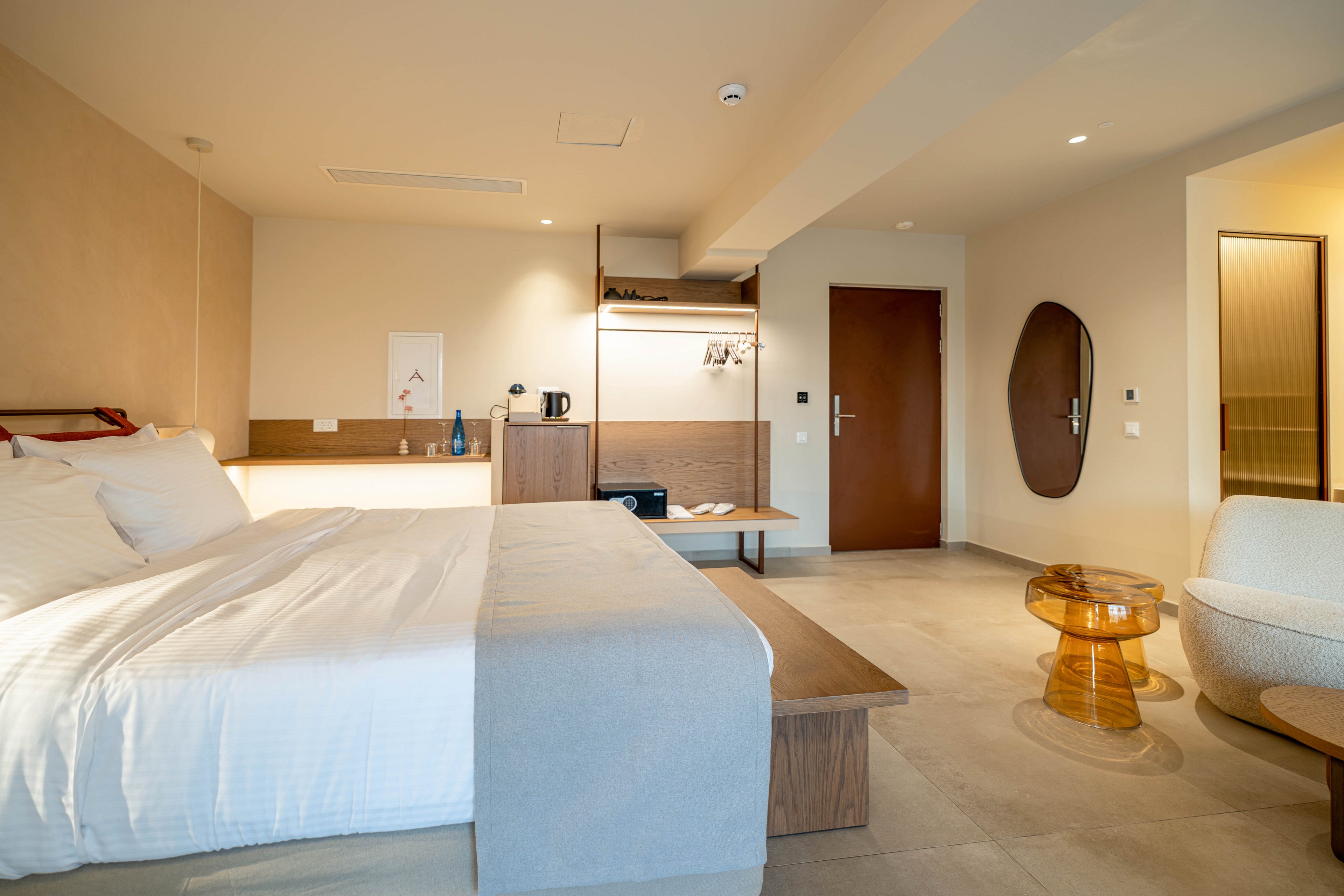 A spacious modern hotel room features a large neatly made bed with white linens and a light beige throw. The room has minimalist wooden furniture, including a low bench, open wardrobe, and a small desk area with a kettle and cups. A round amber-colored side table and a curved upholstered chair add contemporary style to the seating area. Warm neutral tones, soft lighting, and clean lines create a calm, inviting atmosphere.