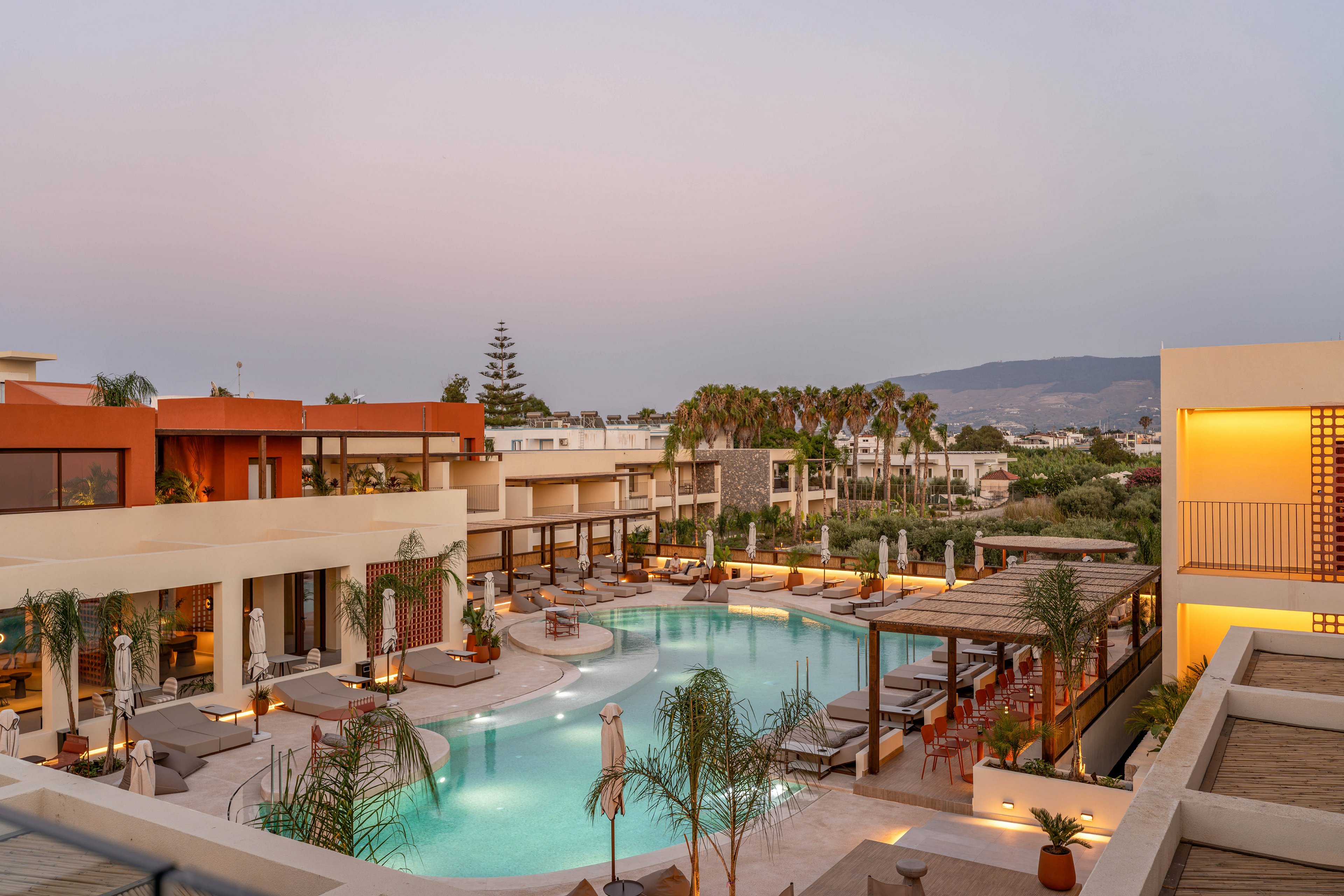 A contemporary resort complex features a large, curved swimming pool surrounded by sun loungers and umbrellas. Warm lights reflect on the water as evening approaches, creating a tranquil atmosphere. The buildings showcase modern architecture with flat roofs, terraces, and earth-toned facades. Palm trees and potted plants add a tropical touch, while distant hills and a soft pastel sky frame the scene.