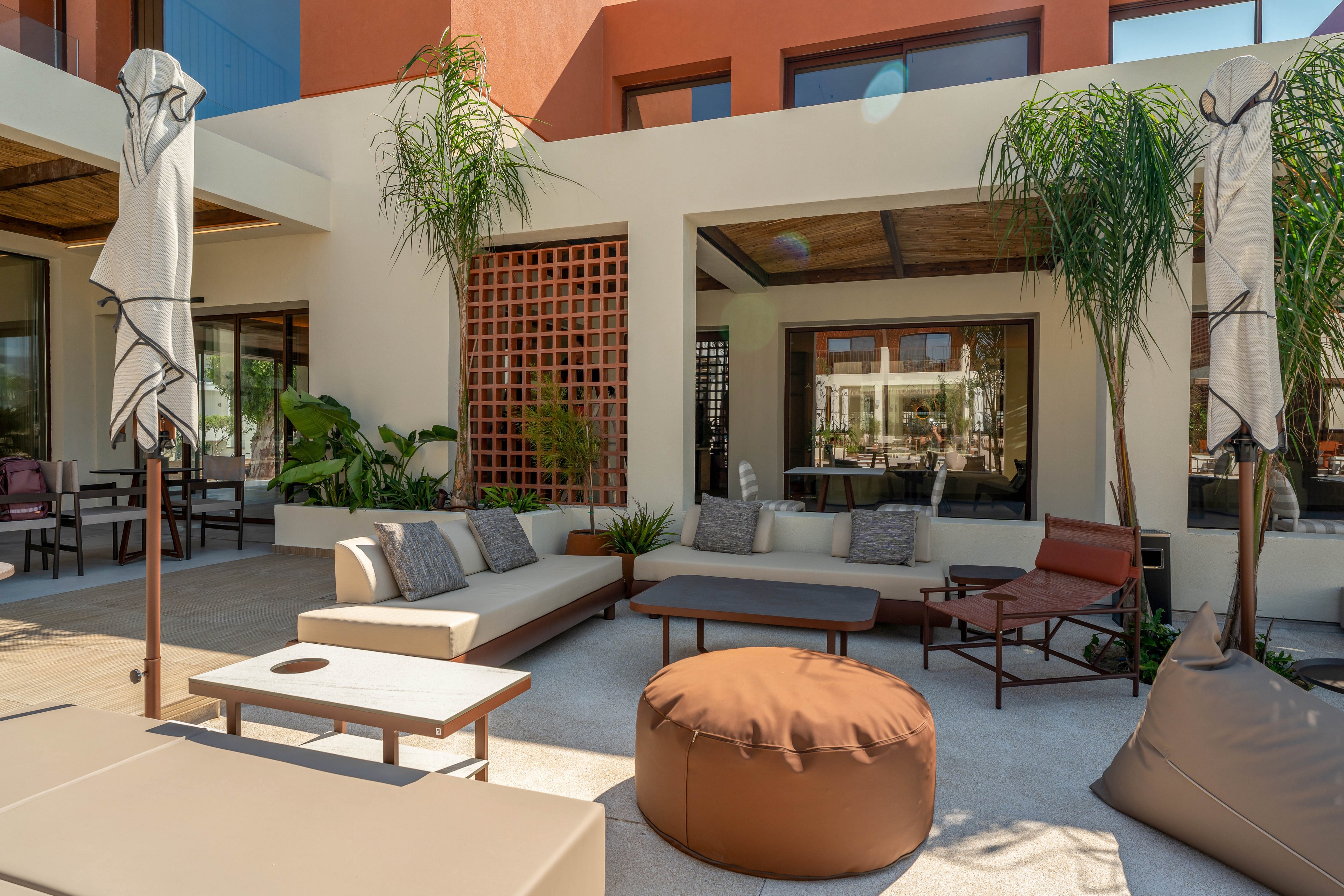 A stylish outdoor lounge area features modern sofas, cushioned chairs, and a round pouf arranged around low tables. The space is set against a contemporary building facade with warm earth-toned walls and large windows. Tall potted palms and other greenery soften the architecture and add a resort-like feel. Neutral fabrics, clean lines, and ample shade create a relaxed, inviting atmosphere for socializing or relaxing.