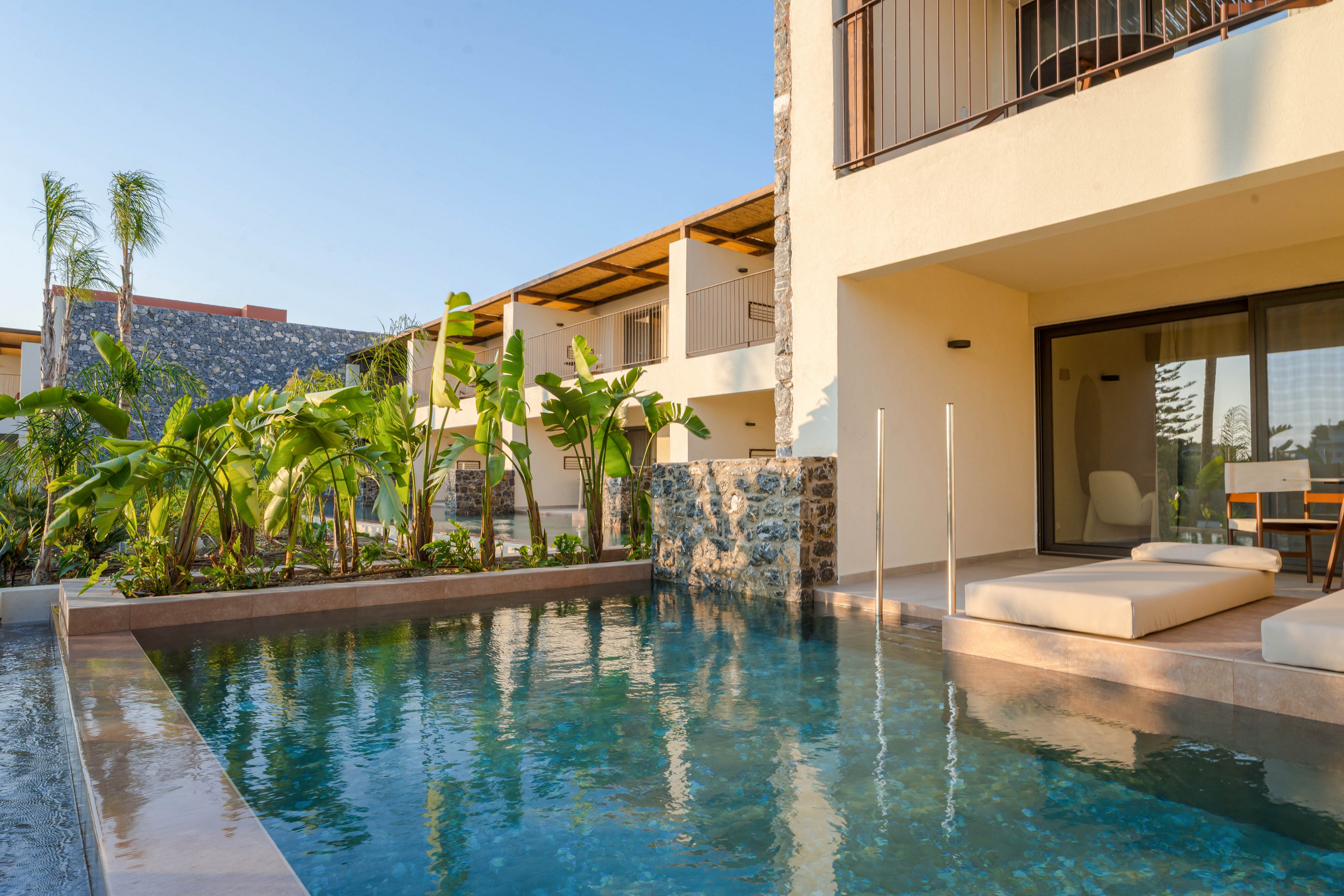 A contemporary resort building features a private pool directly in front of a ground-floor suite. The terrace includes cushioned lounge beds and a small outdoor seating area beside the water. Tropical plants and palm trees line the edge of the pool, adding lush greenery to the scene. Warm sunlight and clear blue sky create a relaxed, vacation atmosphere in this modern outdoor setting.