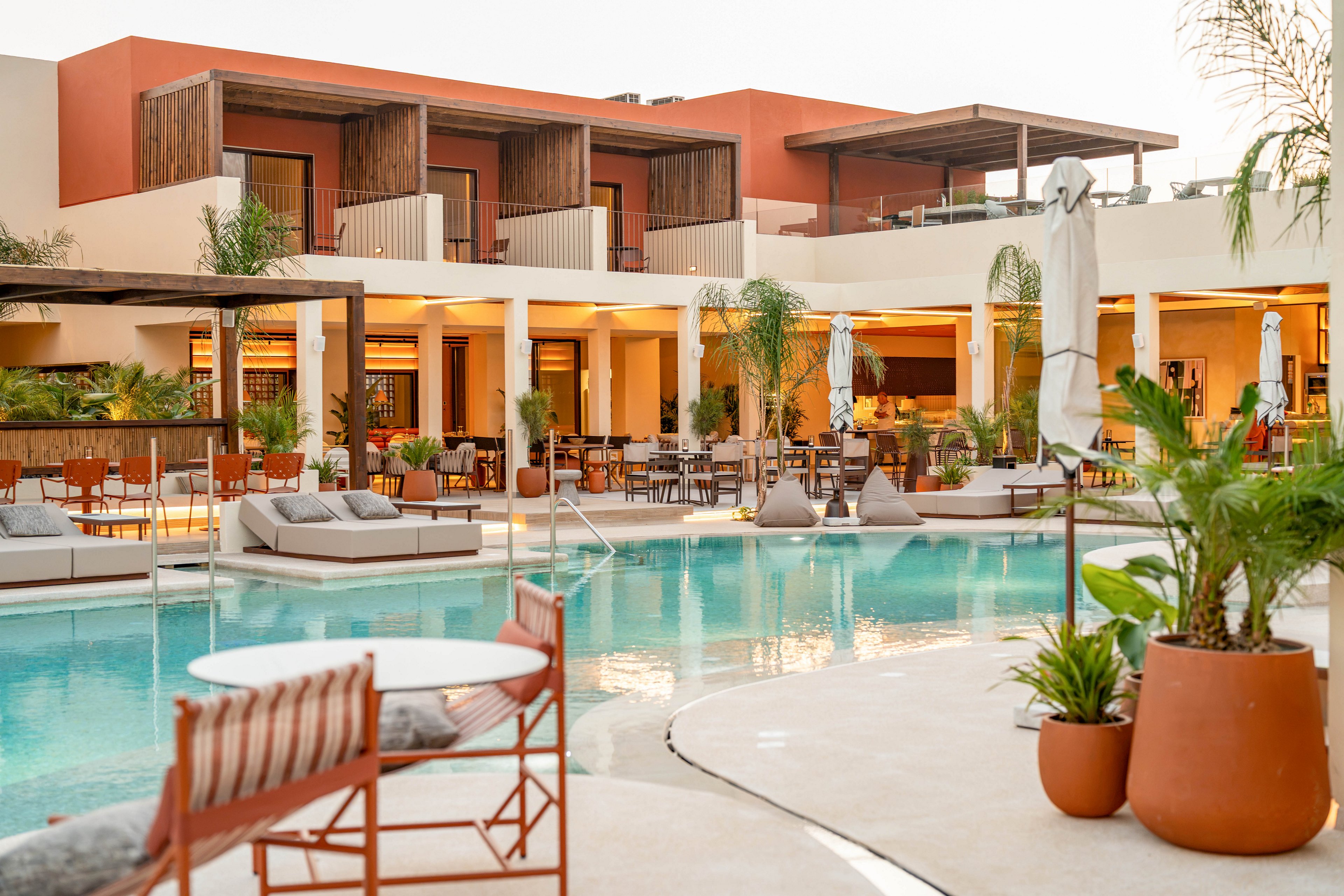 A contemporary resort features a tranquil swimming pool surrounded by lounge chairs and seating areas. Warm-toned buildings with wooden balconies and open-air terraces overlook the water. Potted tropical plants and palm trees add greenery around the pool deck. Soft evening light creates a relaxed, inviting atmosphere for outdoor dining and leisure.